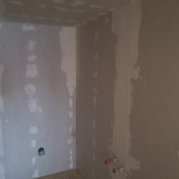 Tynki maszynowe - Narożnik pomieszczenia w trakcie prac wykończeniowych. Ściany pokryte szpachlą i łatami gipsowymi, widoczne instalacje elektryczne i wodne. Przygotowanie do dalszych prac.