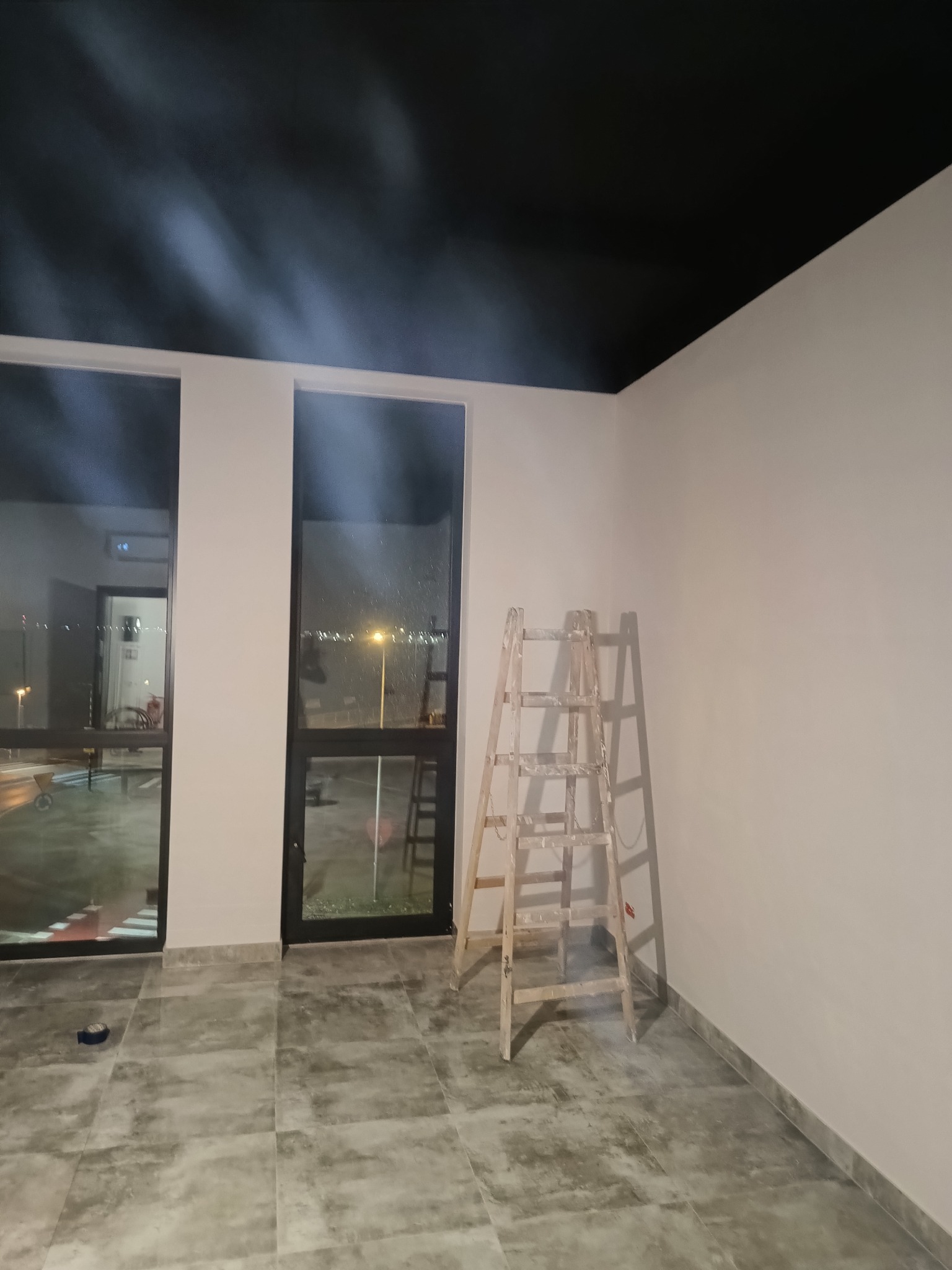 Świeżo pomalowane wnętrze z czarnym sufitem, jasnymi ścianami i drewnianą drabiną. Widoczne okna z nocnym widokiem na zewnątrz. Szare płytki na podłodze.