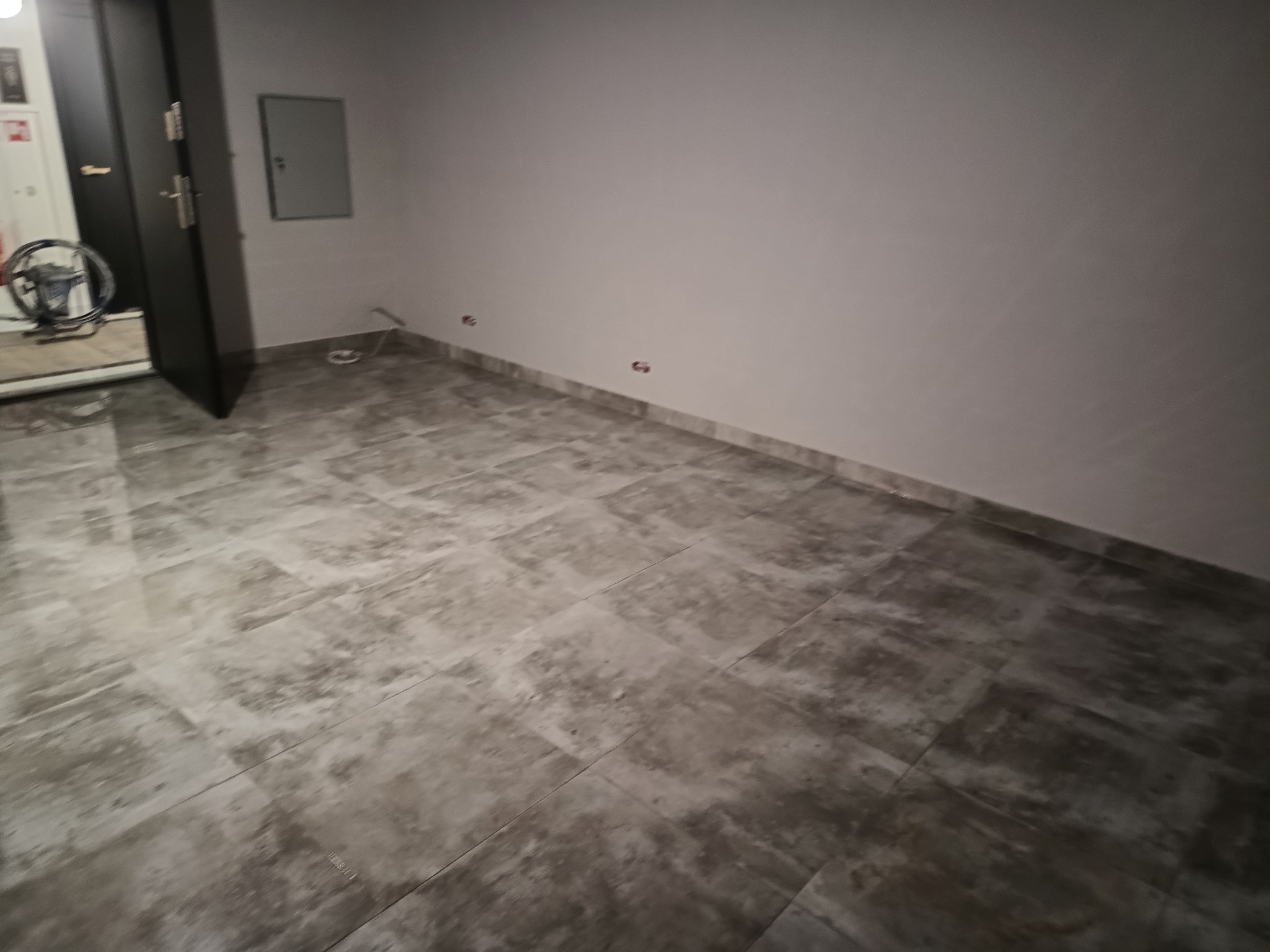 Wykończone wnętrze z szarymi płytkami podłogowymi i świeżo pomalowanymi ścianami. Widoczne punkty elektryczne na ścianie, czarne drzwi i skrzynka elektryczna.