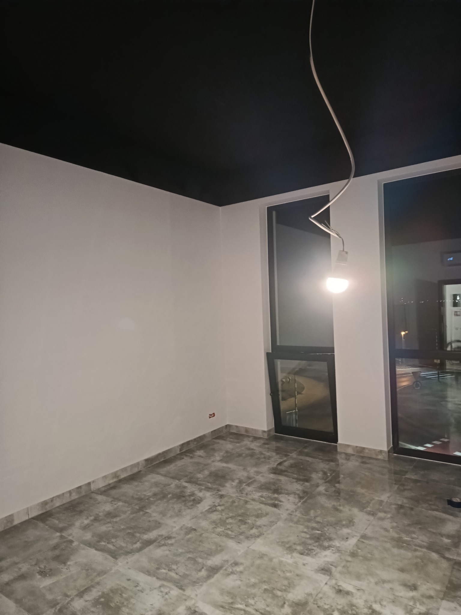 Wykończone wnętrze z czarnym sufitem, białymi ścianami i szarą podłogą z płytek. Widoczne okna z ciemnymi ramami i żarówka na kablu.
