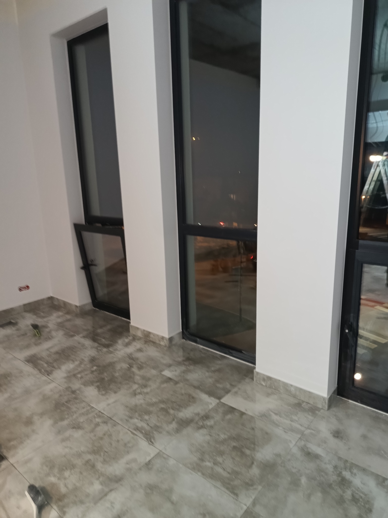 Wykończone wnętrze z dużymi oknami z czarnymi ramami i szarą, błyszczącą podłogą. Widoczne narzędzia malarskie sugerują prace wykończeniowe.