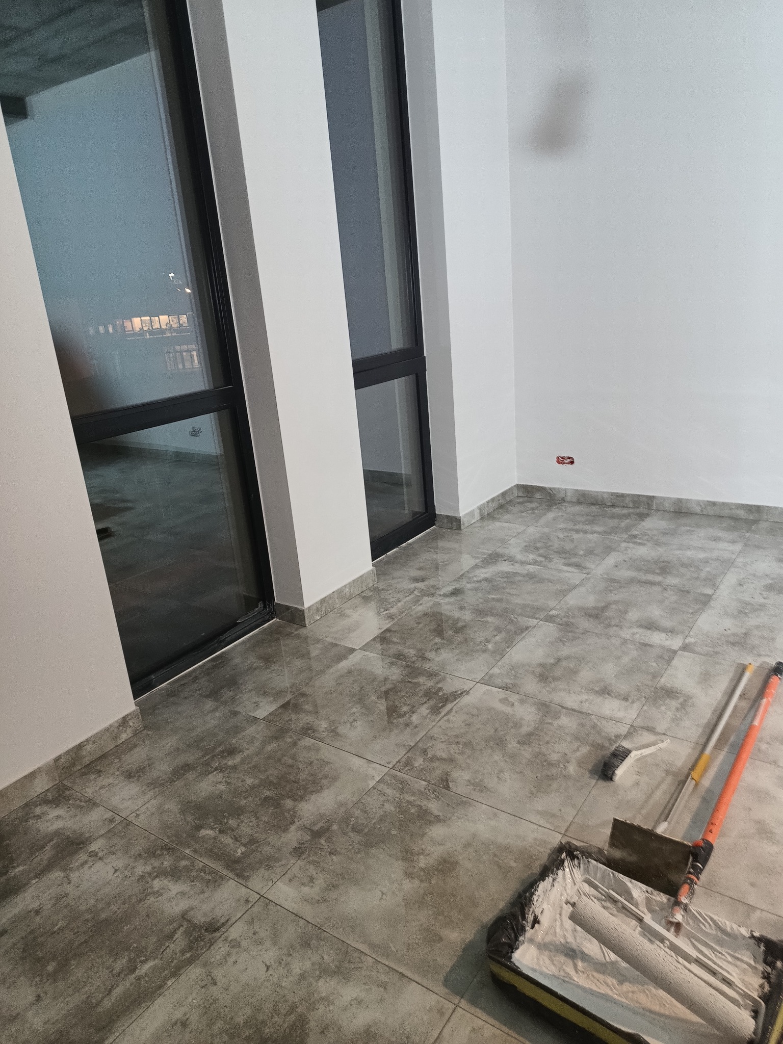 Pokój po malowaniu ścian na biało, widoczne narzędzia malarskie: wałek w kuwecie, pędzel i teleskopowy kij. Szare płytki na podłodze, duże okna.