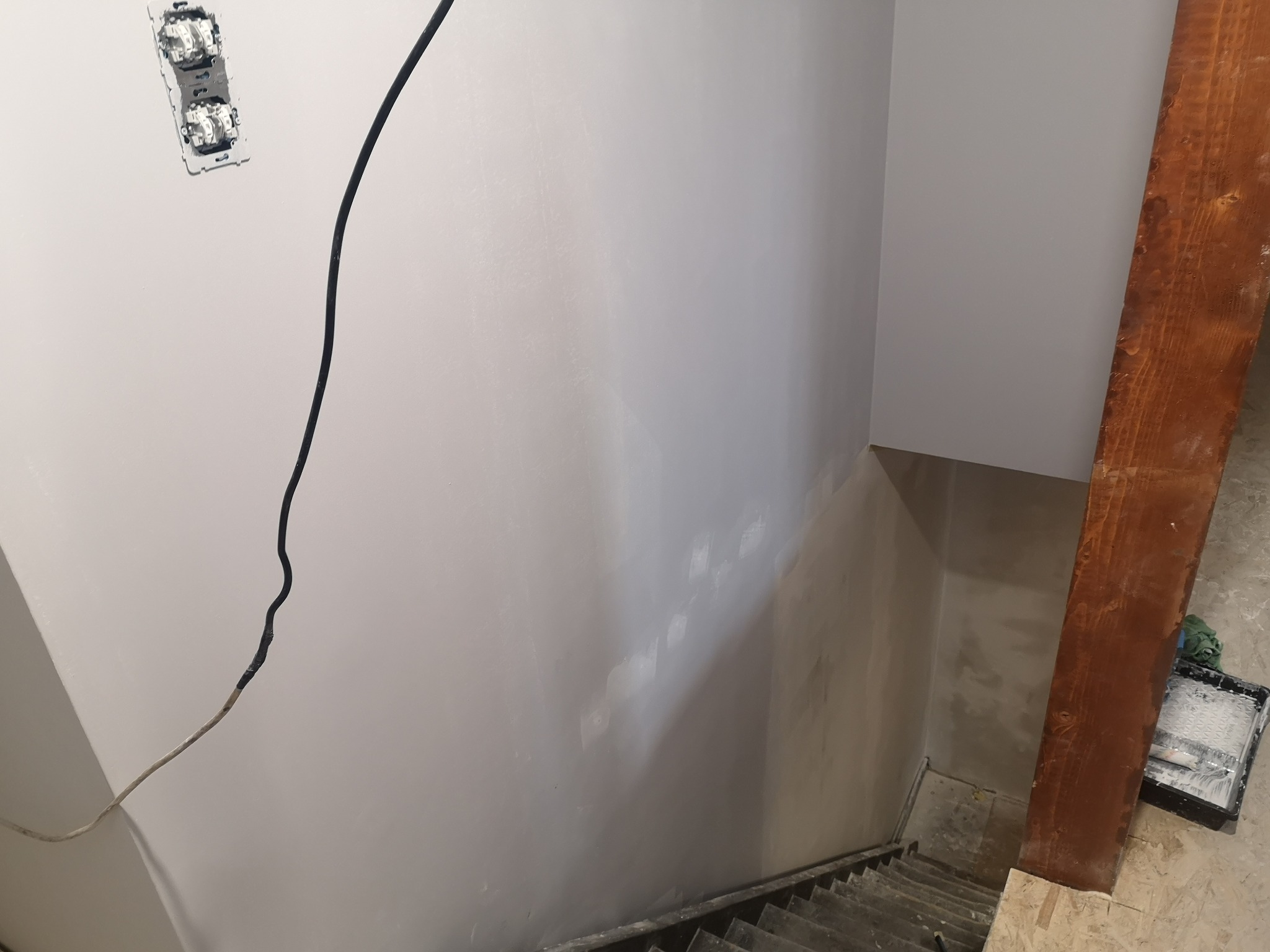 Świeżo pomalowana ściana schodów z widoczną puszką elektryczną i kablem. W tle schody i fragment drewnianej belki. Widoczny ślad po łataniu ubytków.