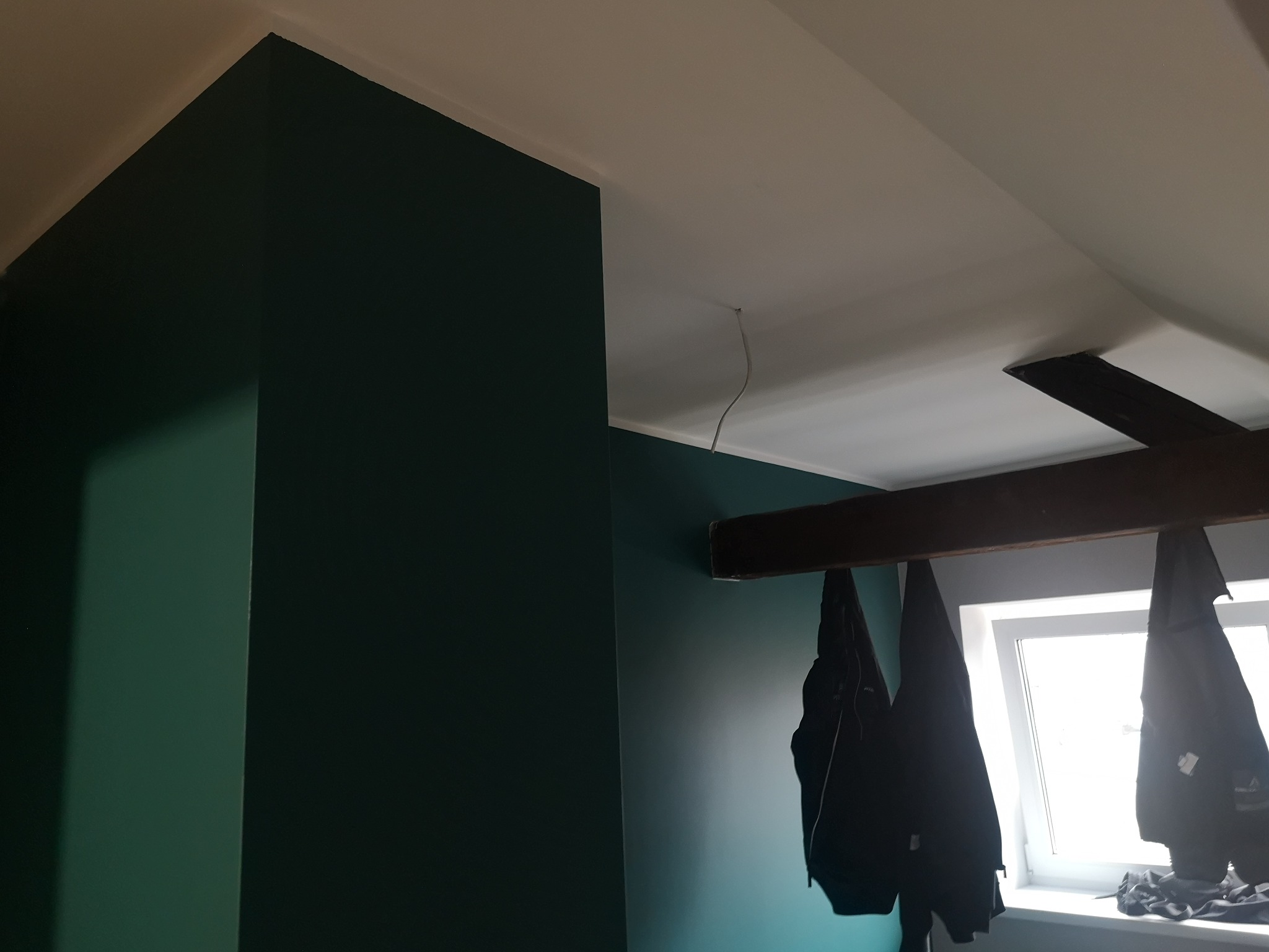 Ściana pomalowana na ciemny zielony kolor w pokoju na poddaszu z oknem i widoczną belką stropową. Na belce wiszą kurtki.