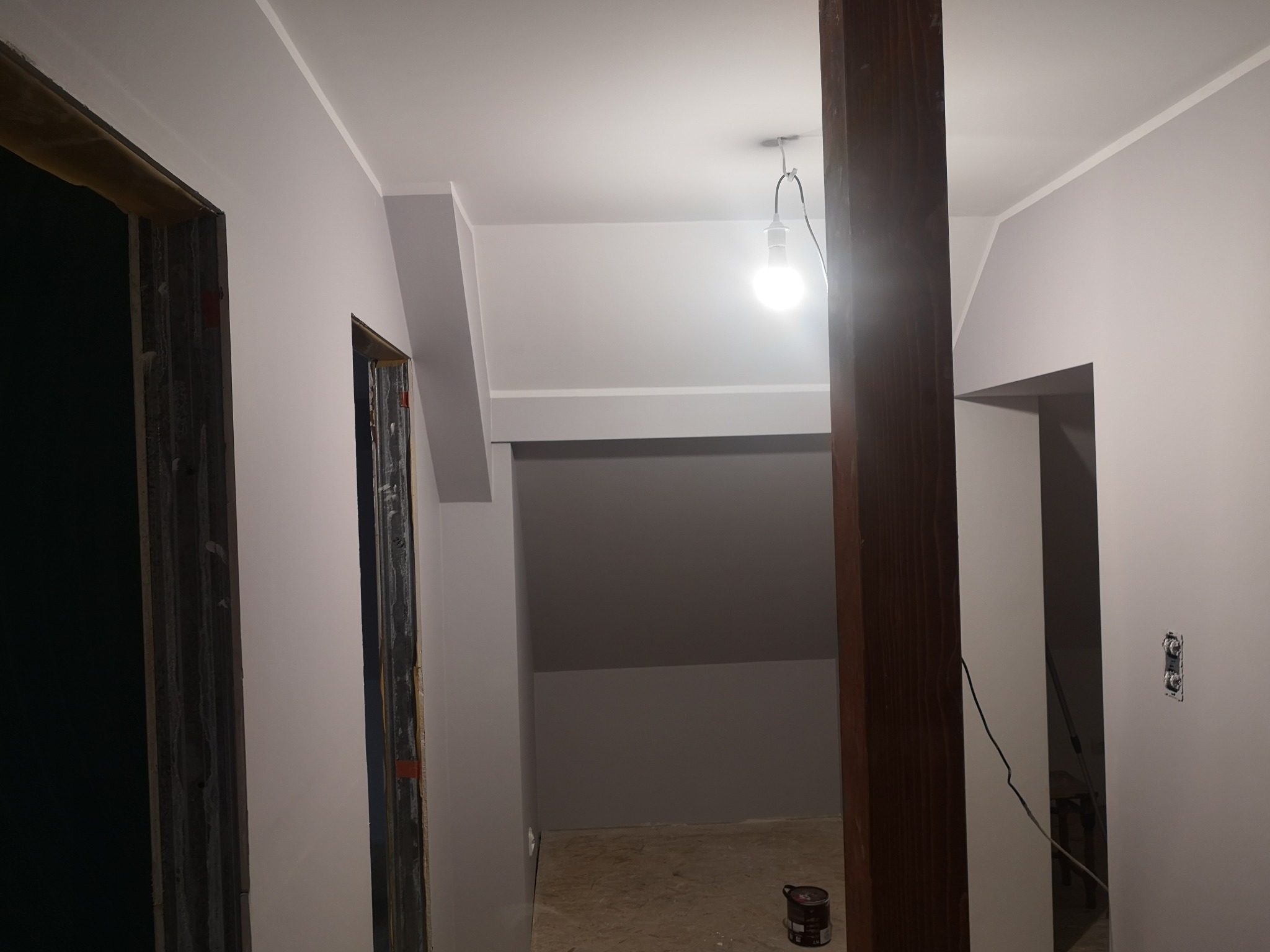 Wnętrze po malowaniu: jasne ściany w odcieniach szarości, drewniany słup, widoczne ościeżnice drzwiowe i tymczasowe oświetlenie z żarówką. Puszka z farbą przy słupie.