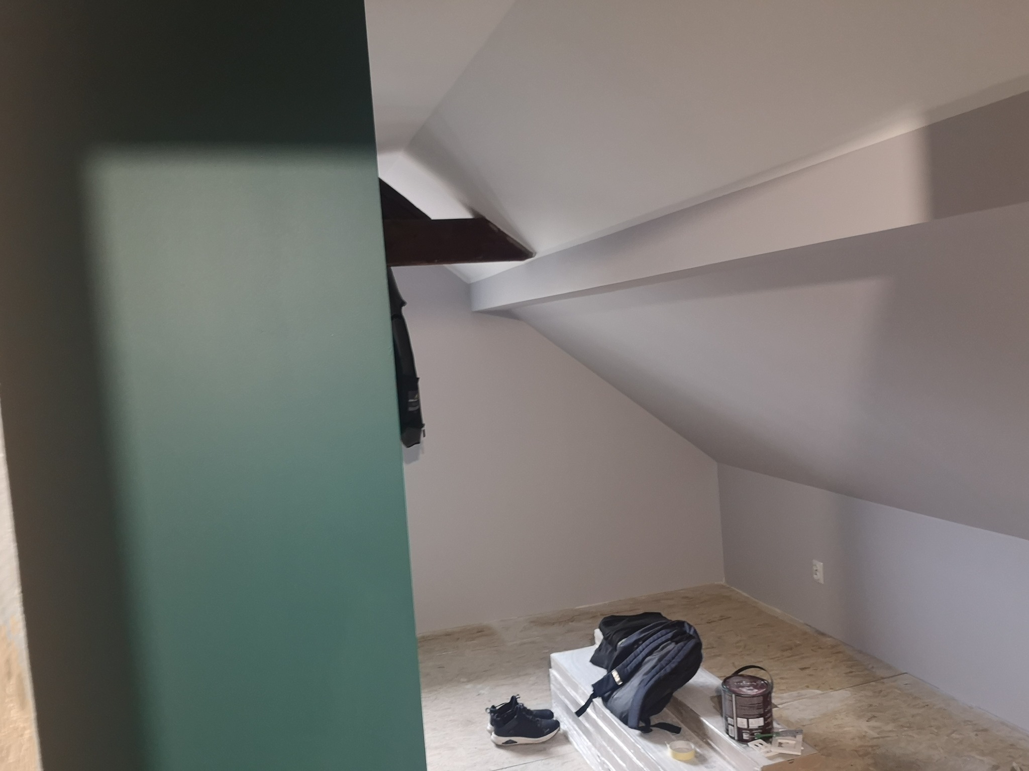 Pomalowane poddasze ze skosami w Czempiniu. Widoczna puszka farby, buty i plecak. Ściany szare, fragment ściany w kolorze butelkowej zieleni. Oświetlenie naturalne.
