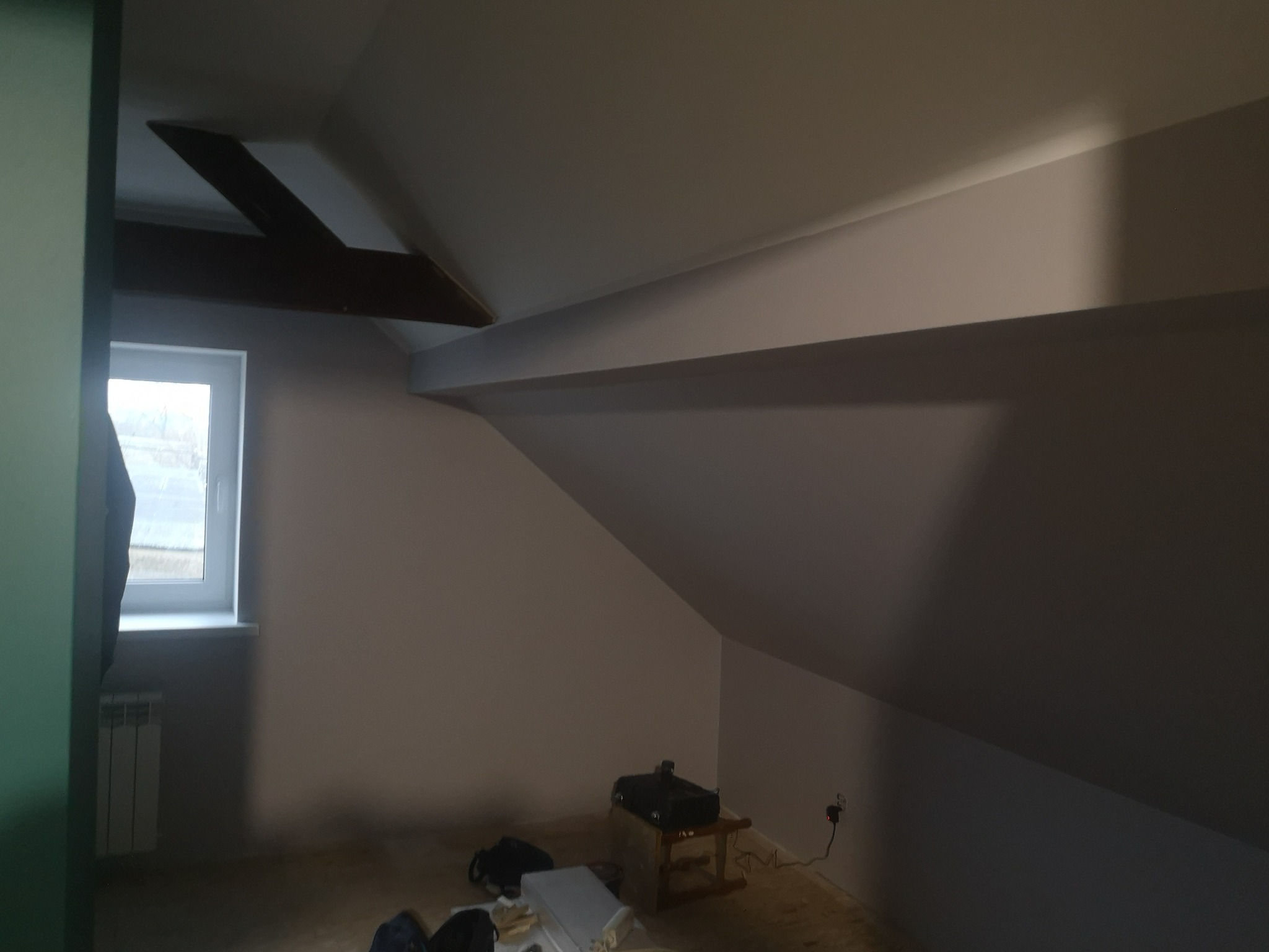 Pokój na poddaszu po malowaniu ścian na jasny odcień szarości, z widocznym oknem, grzejnikiem i drewnianą belką stropową.