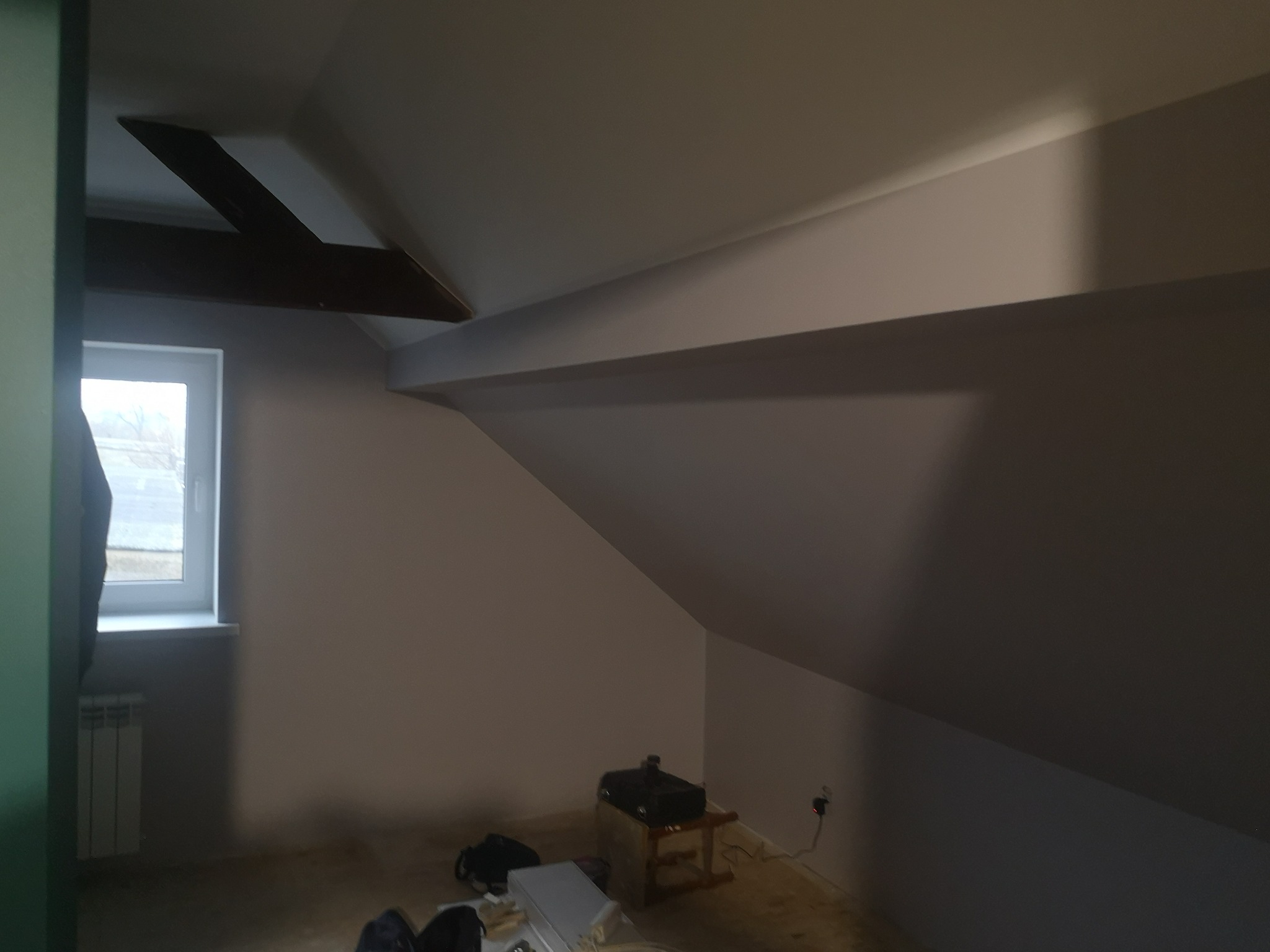 Poddasze po malowaniu na jasny kolor, widoczne skośne ściany, okno z grzejnikiem i drewniana belka stropowa. Na podłodze leżą przedmioty, w rogu stoi projektor.