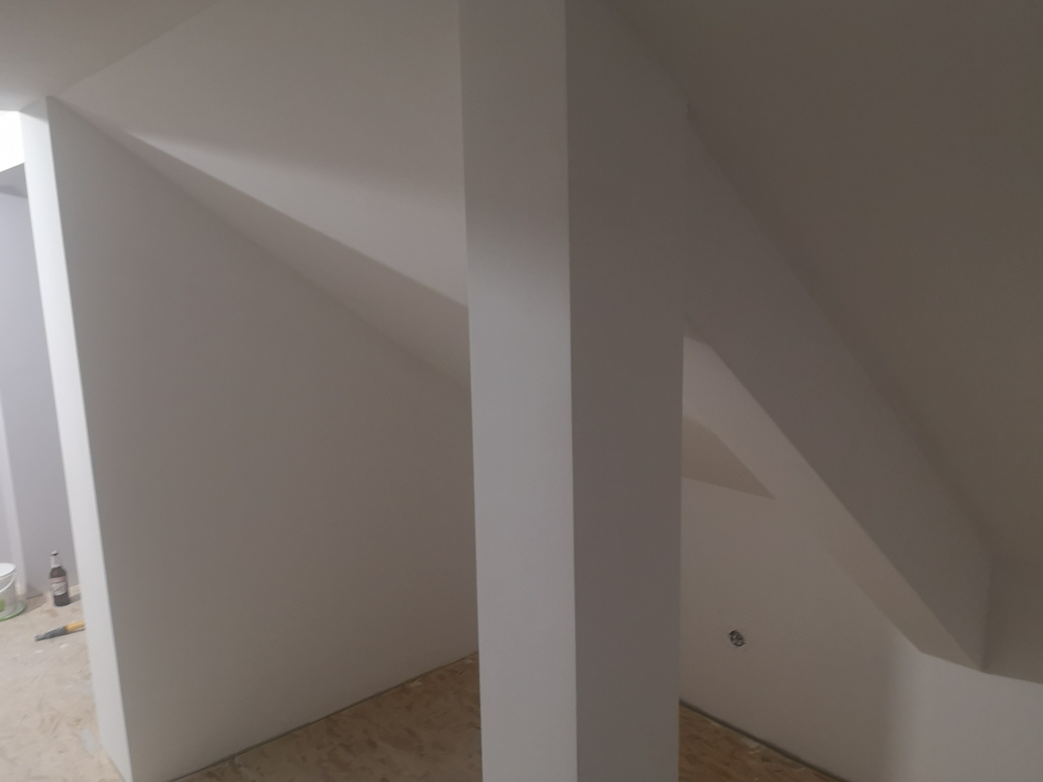 Białe, świeżo pomalowane poddasze ze skośnymi ścianami i filarem. Widoczne ślady po remoncie i butelka. Minimalistyczny design, jasne wnętrze.