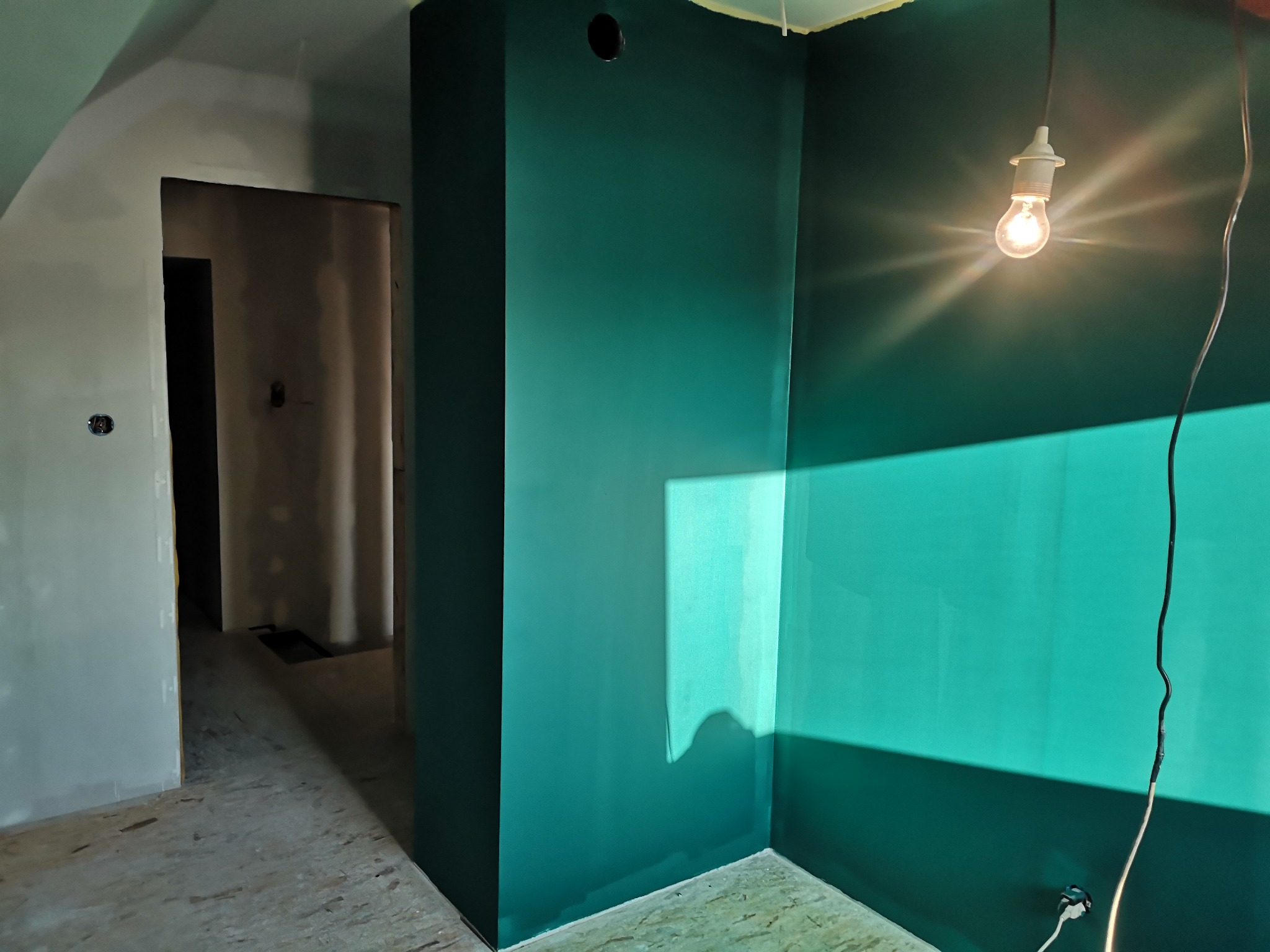 Ściana pomalowana na szmaragdowo w pokoju ze świecącą żarówką. Widoczny fragment sufitu i podłogi w trakcie prac wykończeniowych.