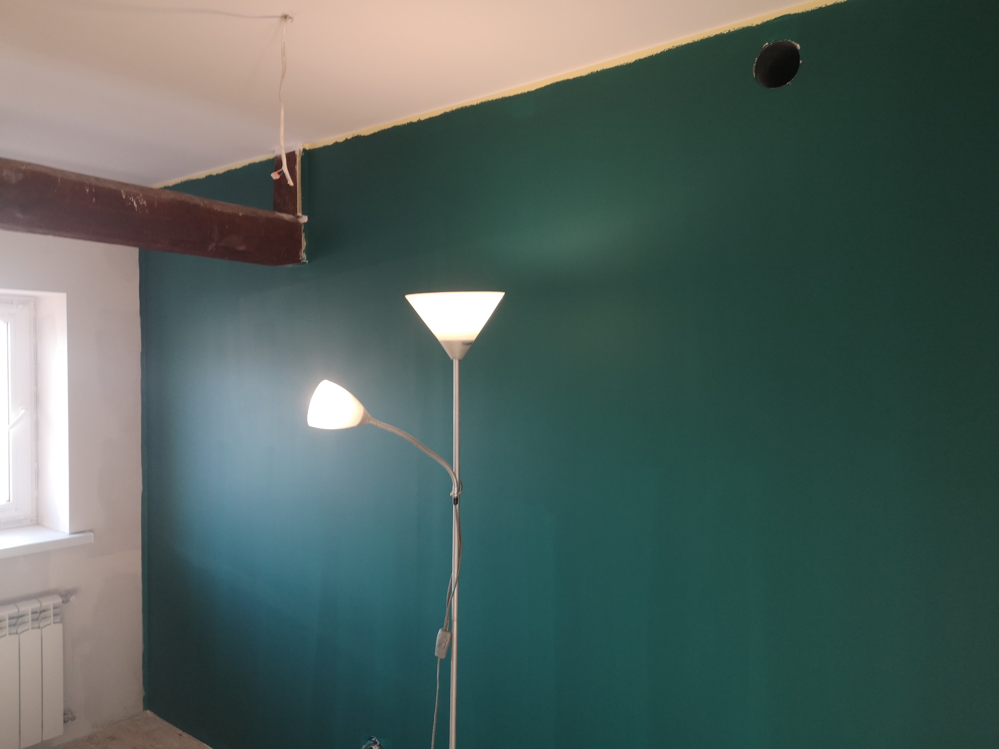 Ściana pomalowana na butelkową zieleń, z widocznym fragmentem drewnianej belki stropowej i lampą podłogową. Przygotowanie do wykończenia wnętrza.
