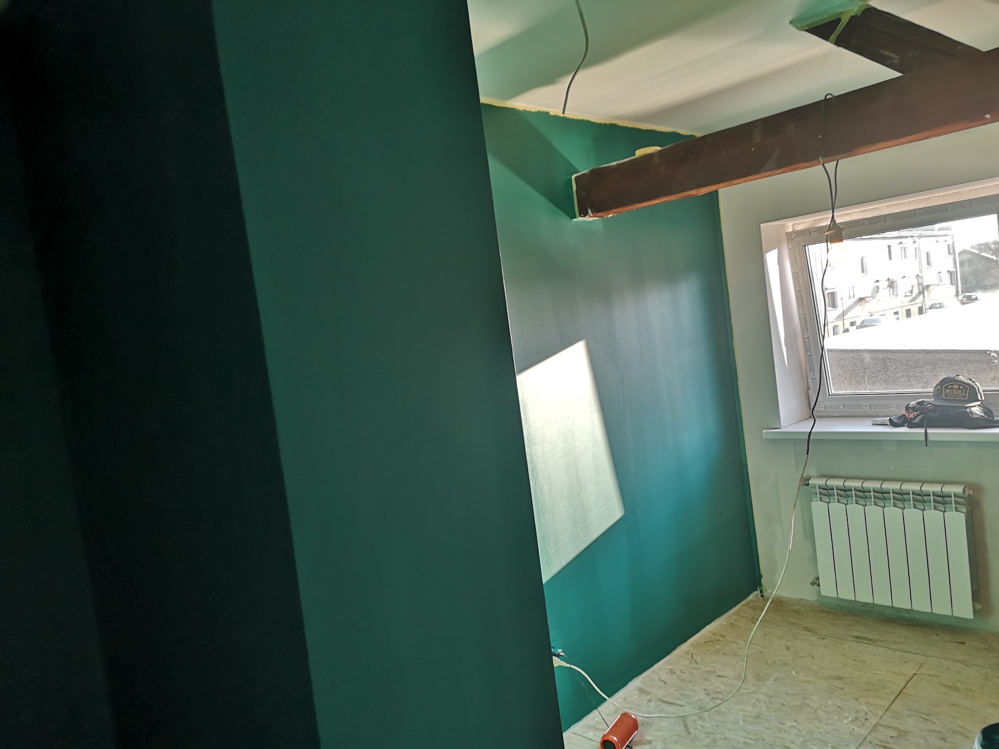 Ściana pomalowana na ciemnozielony kolor w pokoju z oknem. Widoczny grzejnik, surowa podłoga, drewniana belka stropowa i kabel zwisający z sufitu.