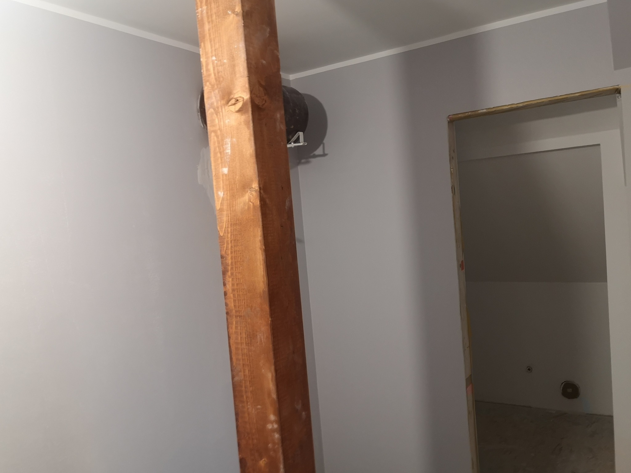 Szara ściana z drewnianą belką i otworem wentylacyjnym. Widoczny fragment ościeżnicy i wnętrze pomieszczenia w trakcie prac wykończeniowych.