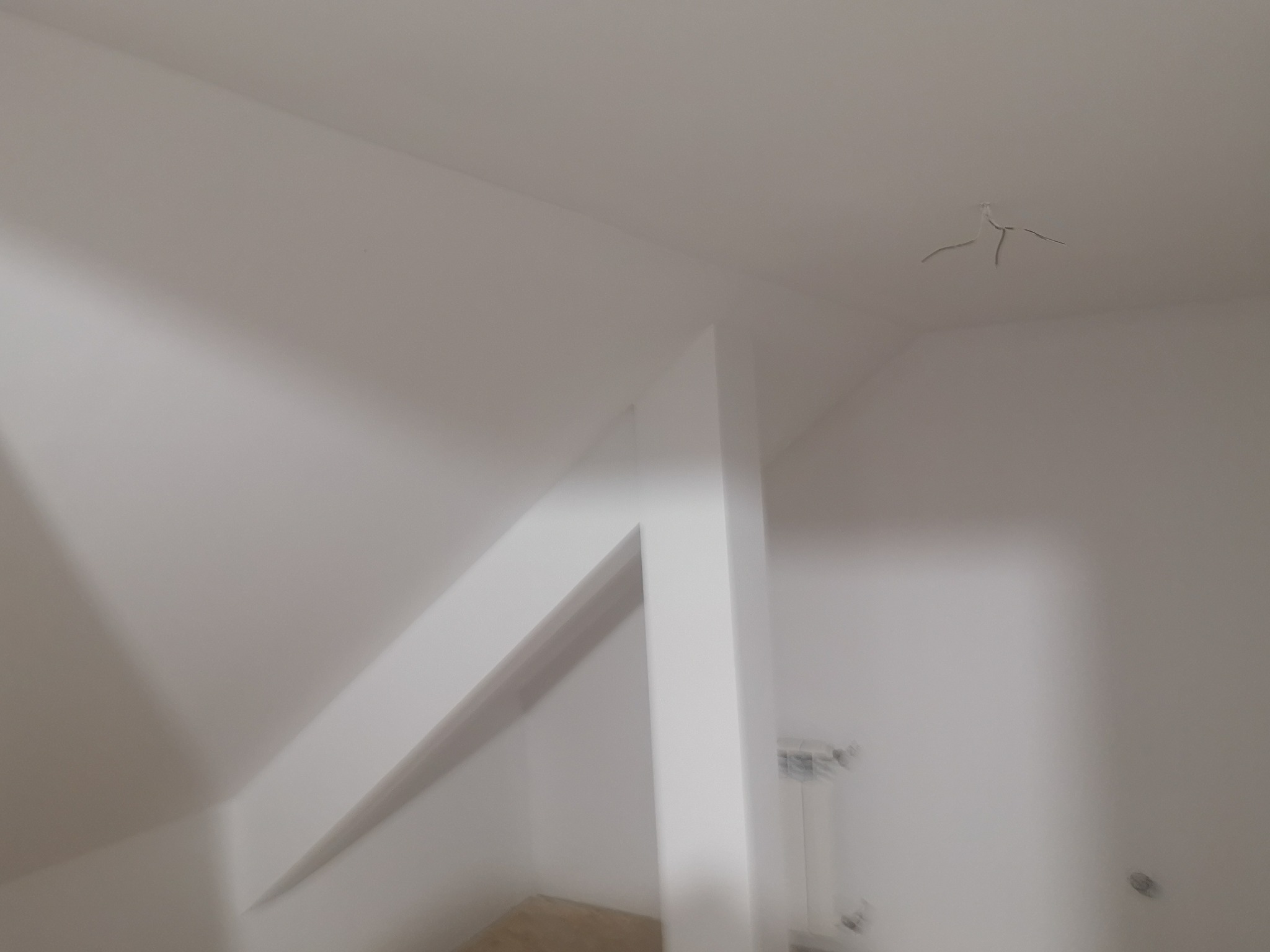 Białe wnętrze poddasza po remoncie, widoczne gładkie ściany, fragment grzejnika i wystające kable z sufitu. Kadrowanie od dołu, z lekkim rozmytym efektem.