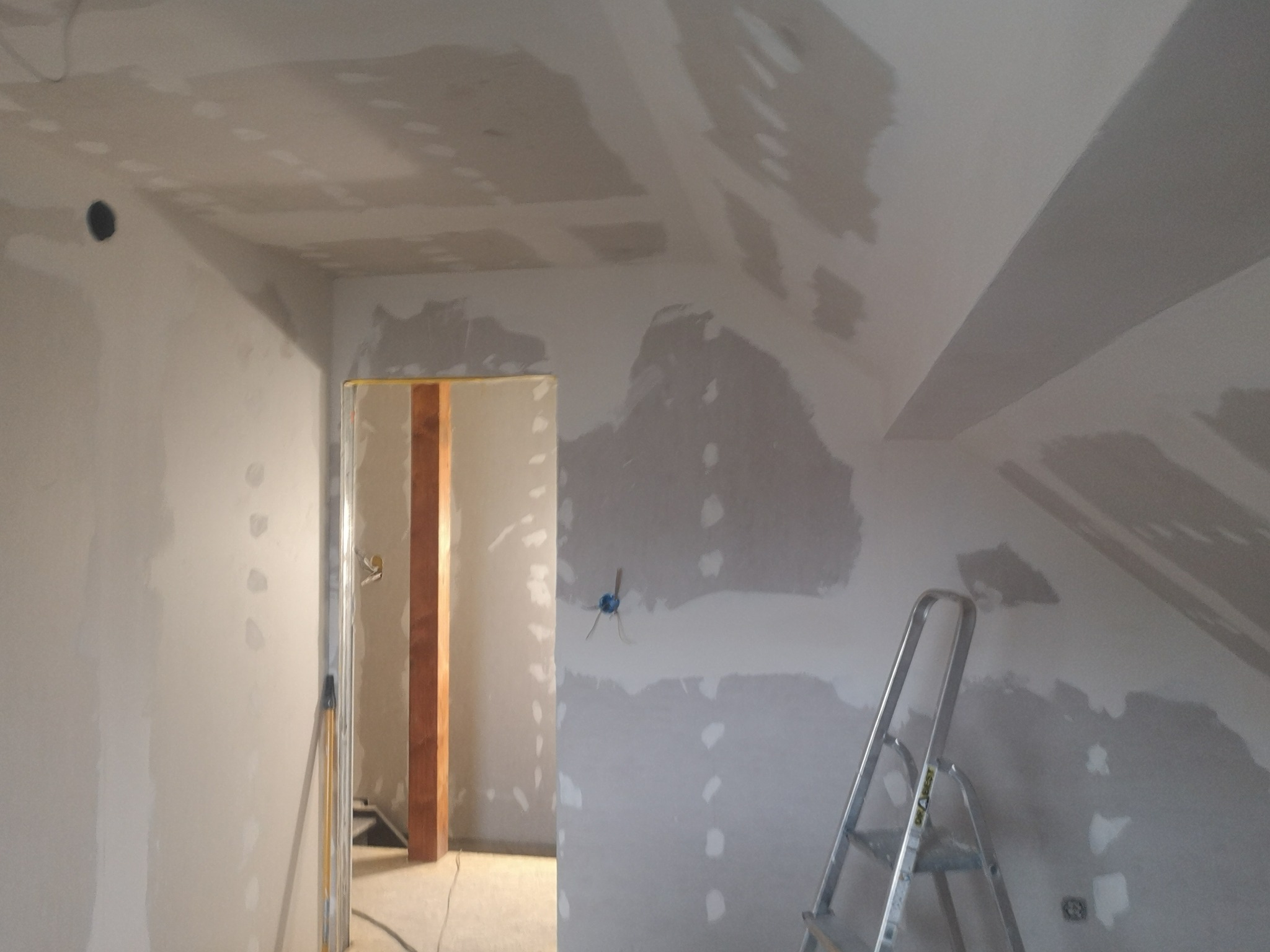 Poddasze w trakcie prac wykończeniowych: ściany i sufit pokryte gładzią szpachlową, widoczna drabina i otwór drzwiowy z drewnianą ościeżnicą.