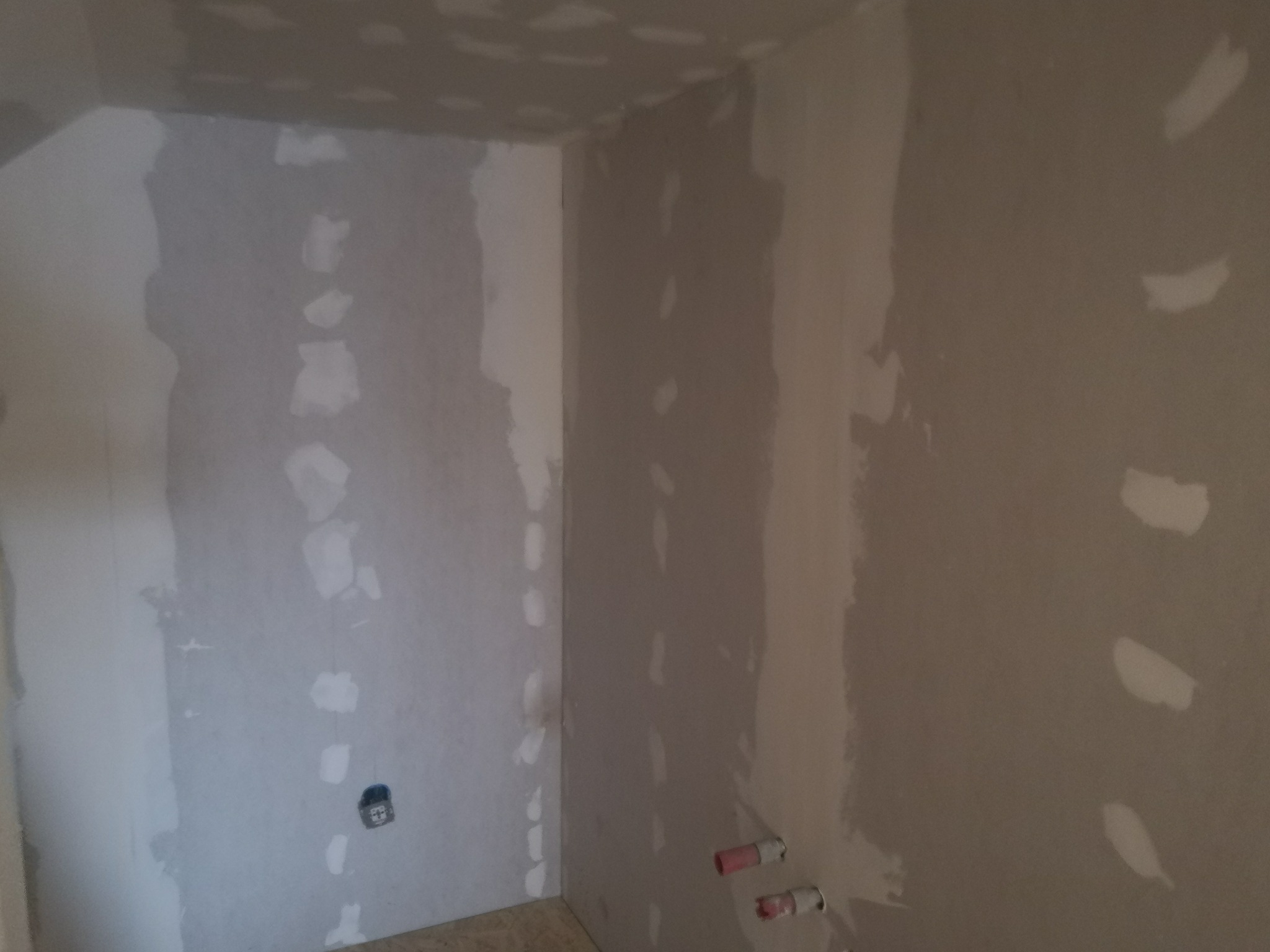 Narożnik pomieszczenia w trakcie prac wykończeniowych. Ściany pokryte szpachlą i łatami gipsowymi, widoczne instalacje elektryczne i wodne. Przygotowanie do dalszych prac.