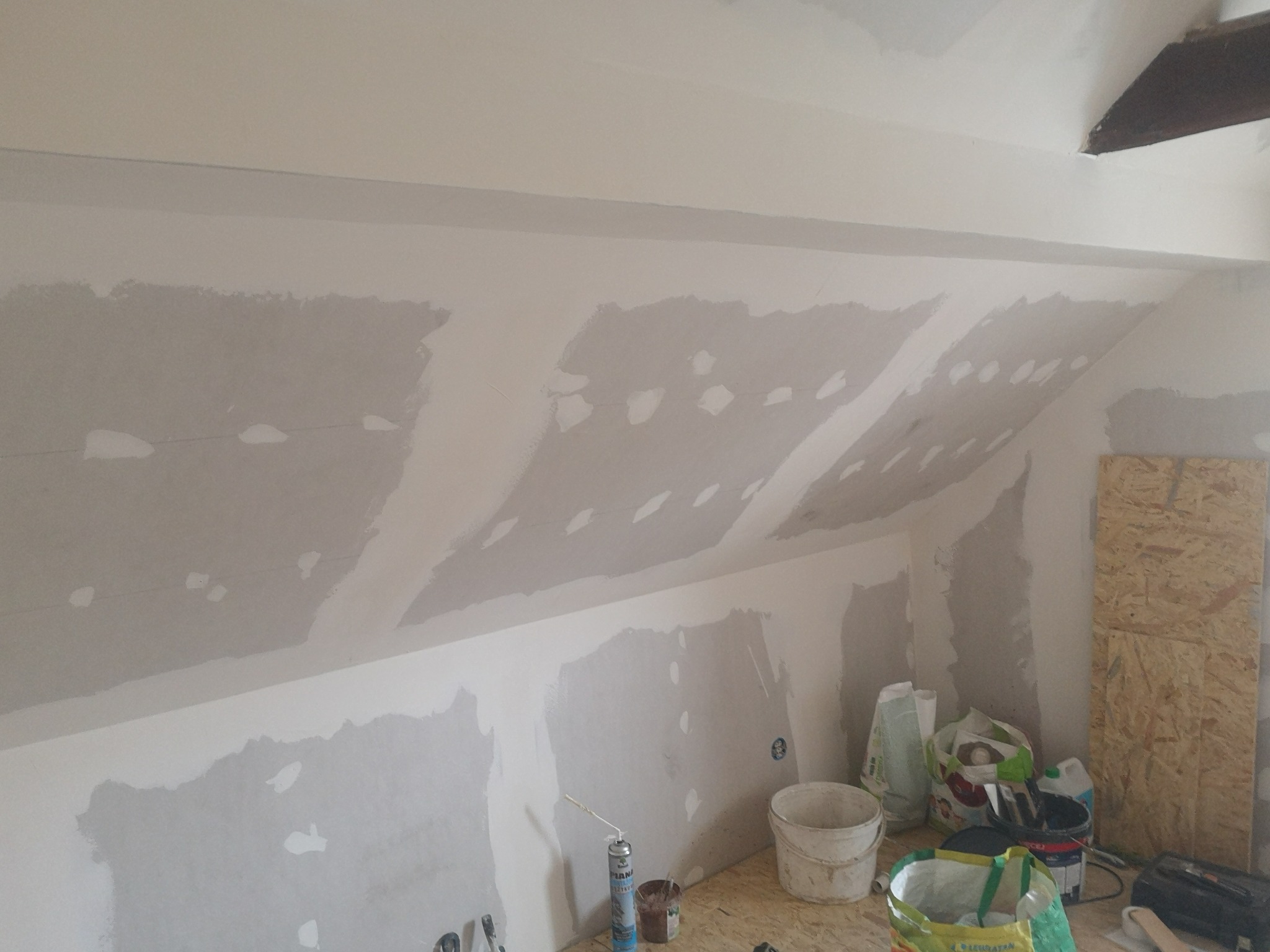 Poddasze w trakcie szpachlowania: widoczne płyty gipsowo-kartonowe na skosach, częściowo pokryte szpachlą. Na podłodze narzędzia i materiały budowlane.