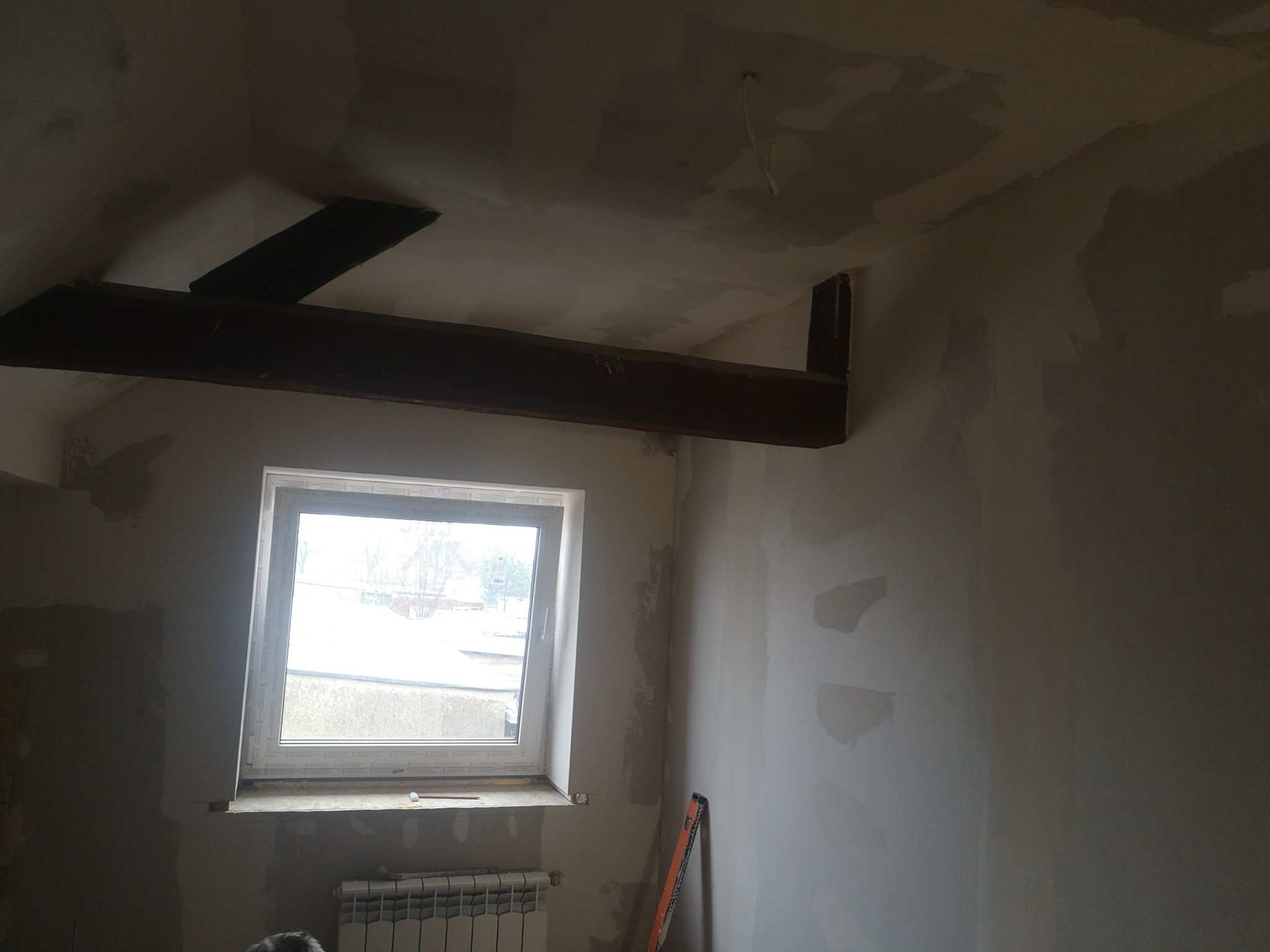 Poddasze w trakcie prac wykończeniowych: ściany i sufit po szpachlowaniu, widoczne drewniane belki stropowe, okno i grzejnik. Poziomica oparta o ścianę.