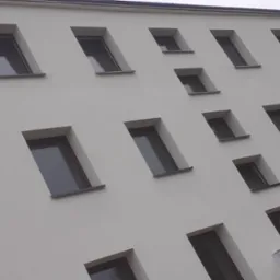 Biała elewacja budynku z regularnie rozmieszczonymi, prostokątnymi oknami z ciemnymi ramami, sfotografowana pod kątem z dołu.