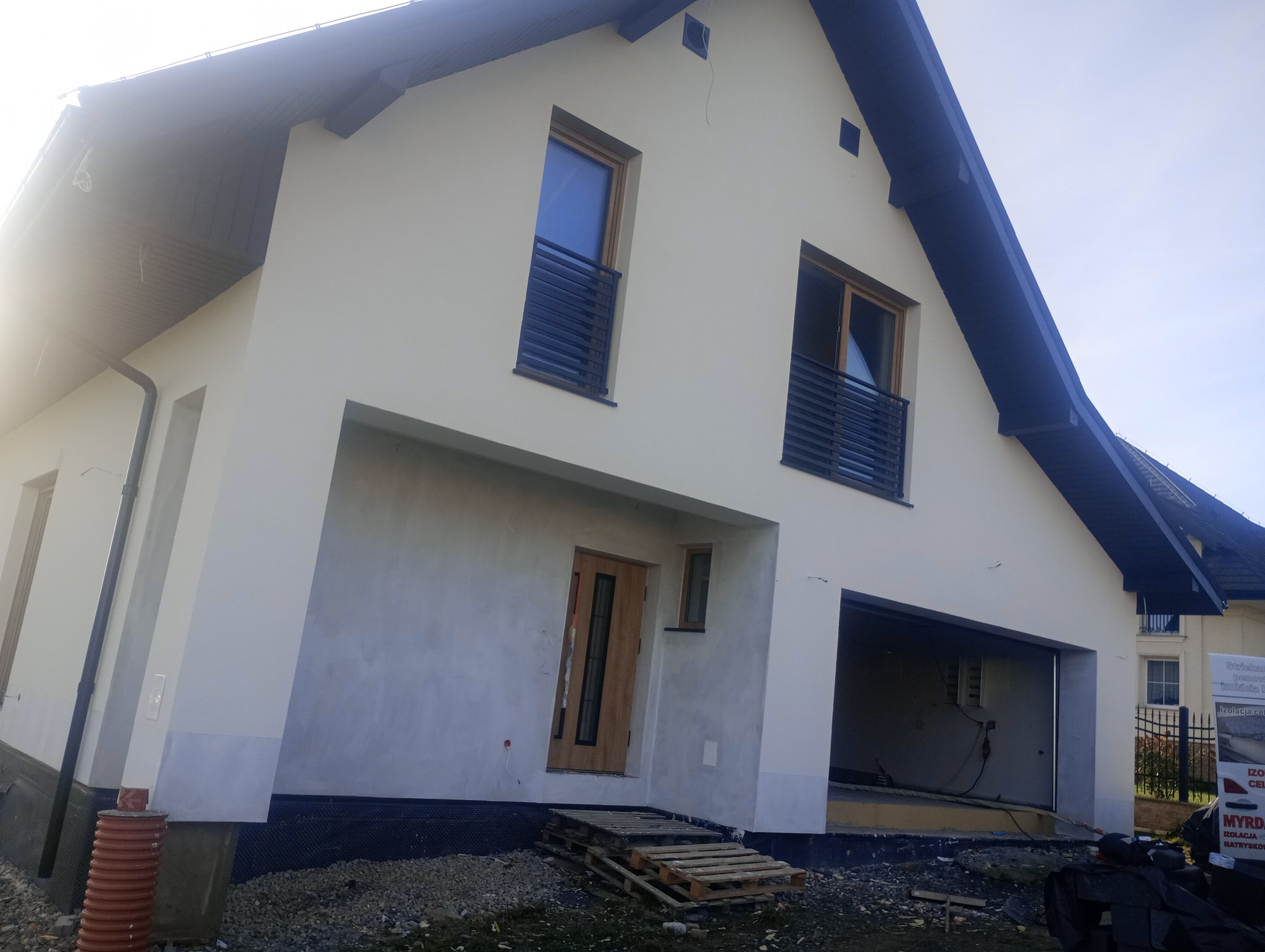 Dom jednorodzinny w Krościenku nad Dunajcem, częściowo pokryty szarą warstwą podkładową, z drewnianymi oknami i drzwiami; widoczne palety przed wejściem i przygotowania do prac wykończeniowych...