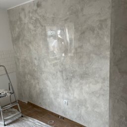 FHU Pro-remont Marcin Jarzębiński - Ściana w szarościach z efektem dekoracyjnym, zabezpieczona podłoga, drabina w rogu. Widoczny kontakt elektryczny i listwa przypodłogowa. Wykończenie wnętrza w trakcie.