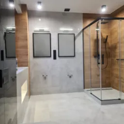 Wykończona łazienka z drewnopodobnymi płytkami w kabinie prysznicowej typu walk-in, szare płytki na ścianie i podłodze, minimalistyczne lustra w czarnych ramach.