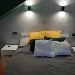 Nowoczesna sypialnia z szarą ścianą imitującą beton, oświetlona lampami ściennymi. Na łóżku żółte, zielone i białe poduszki. Widoczne białe szafki nocne.