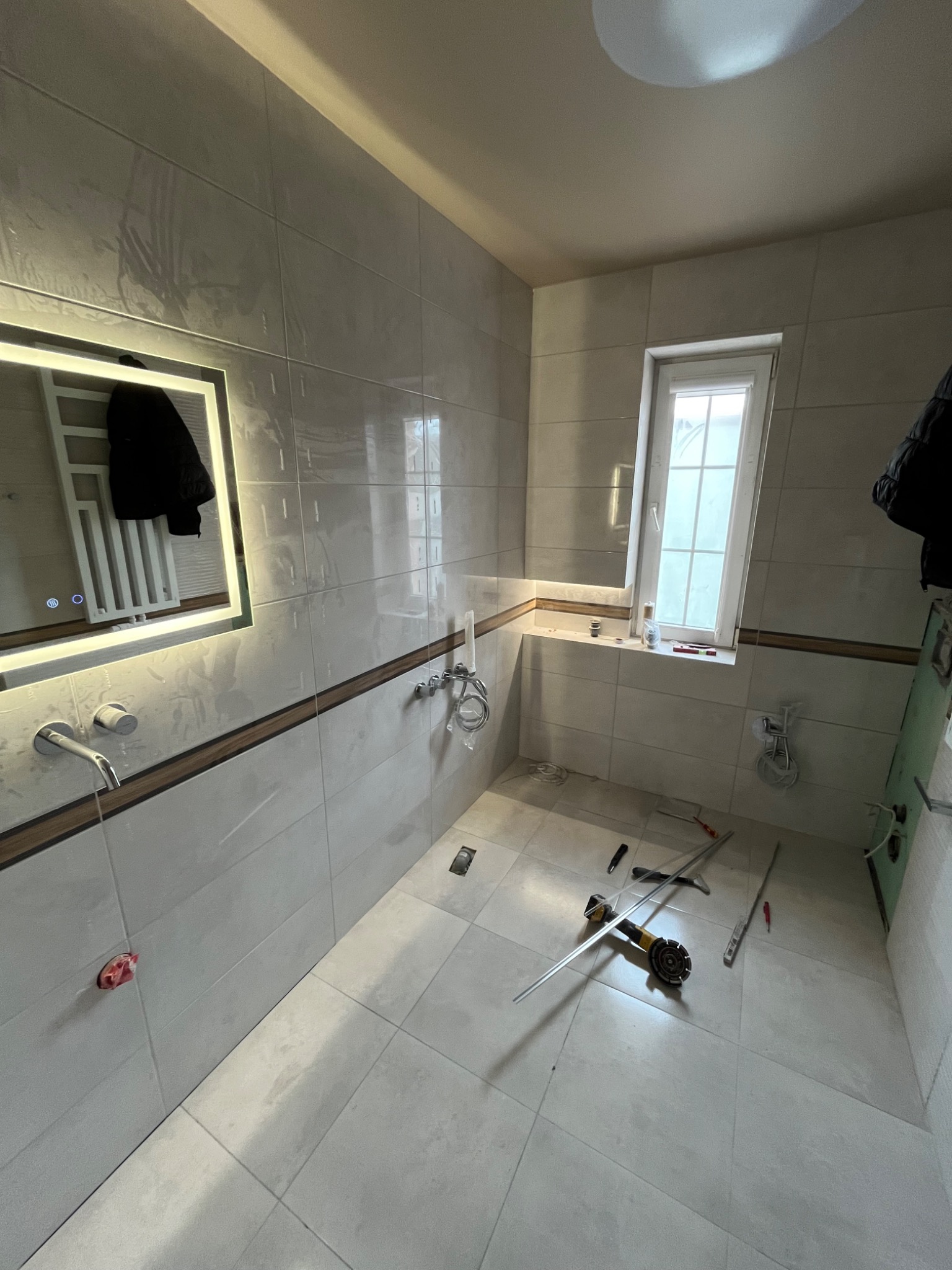 Wnętrze łazienki w trakcie remontu. Widoczne płytki na ścianach i podłodze, lustro z podświetleniem, okno oraz narzędzia glazurnicze na podłodze.