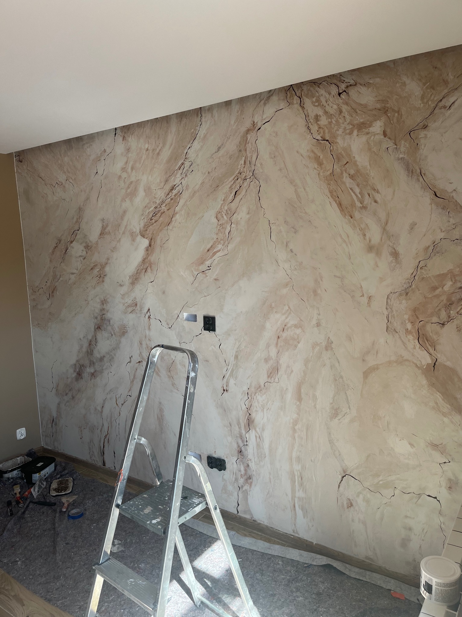 Ściana z dekoracyjną, marmurkową fakturą w odcieniach beżu i brązu. Widoczna drabina malarska i akcesoria malarskie. Dwa gniazdka elektryczne na ścianie.