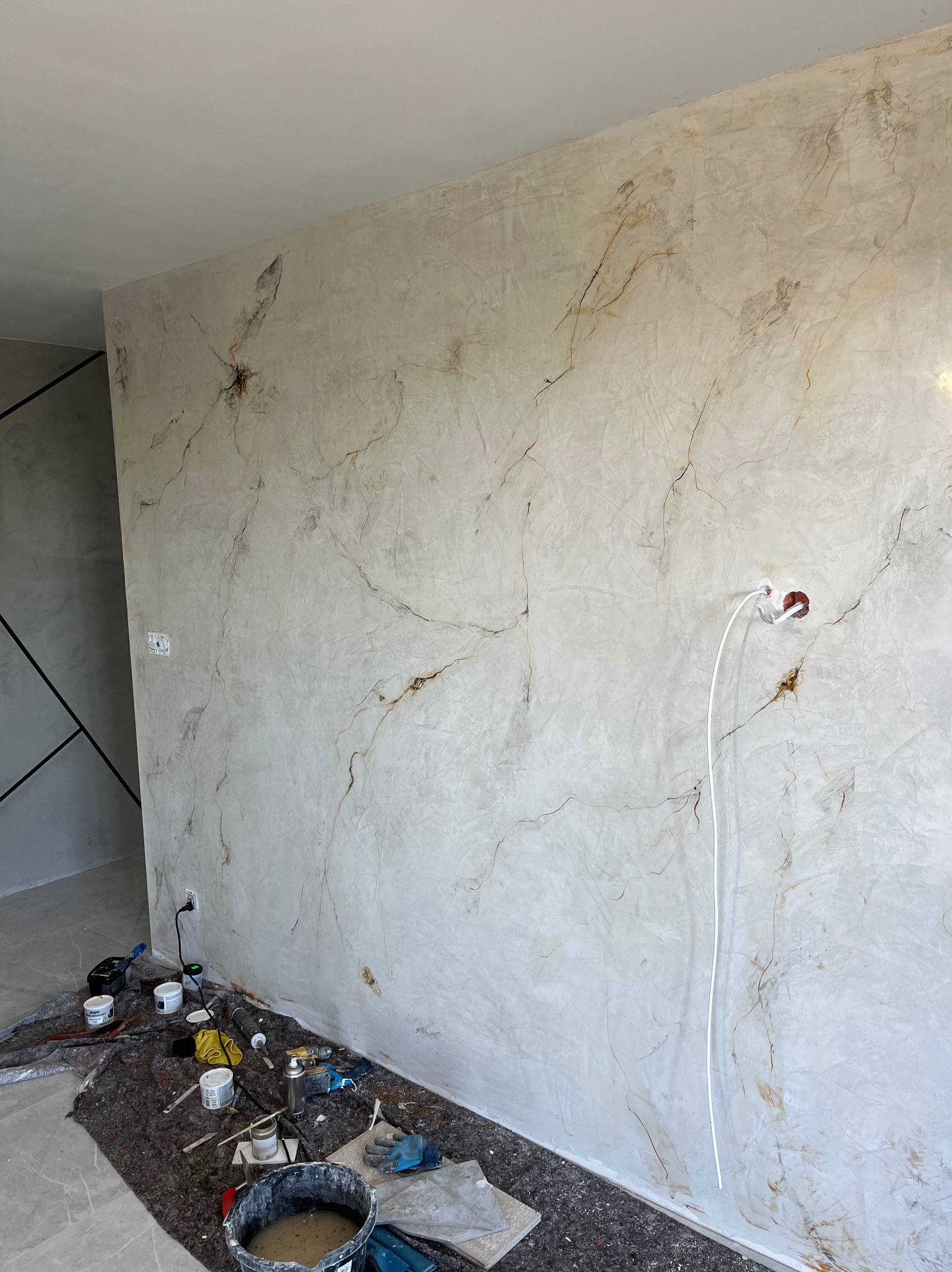 Ściana z efektem marmuru w trakcie realizacji. Widoczne narzędzia malarskie i puszki z farbą. Gniazdko elektryczne wystaje ze ściany.