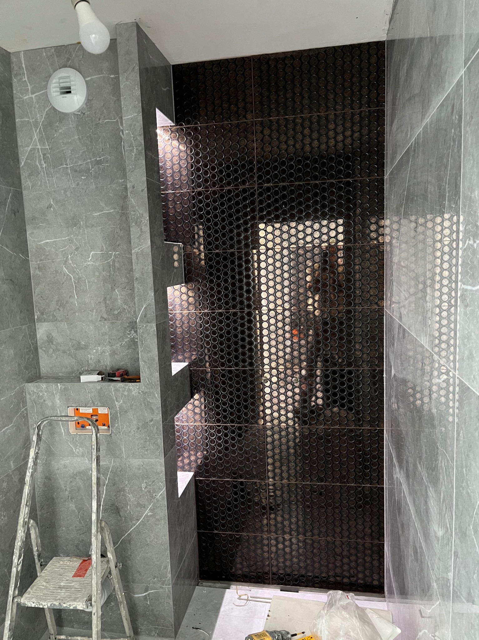 Łazienka w trakcie remontu: szare płytki imitujące marmur, mozaika z małych, okrągłych płytek w strefie prysznica, drabina malarska i narzędzia glazurnicze.