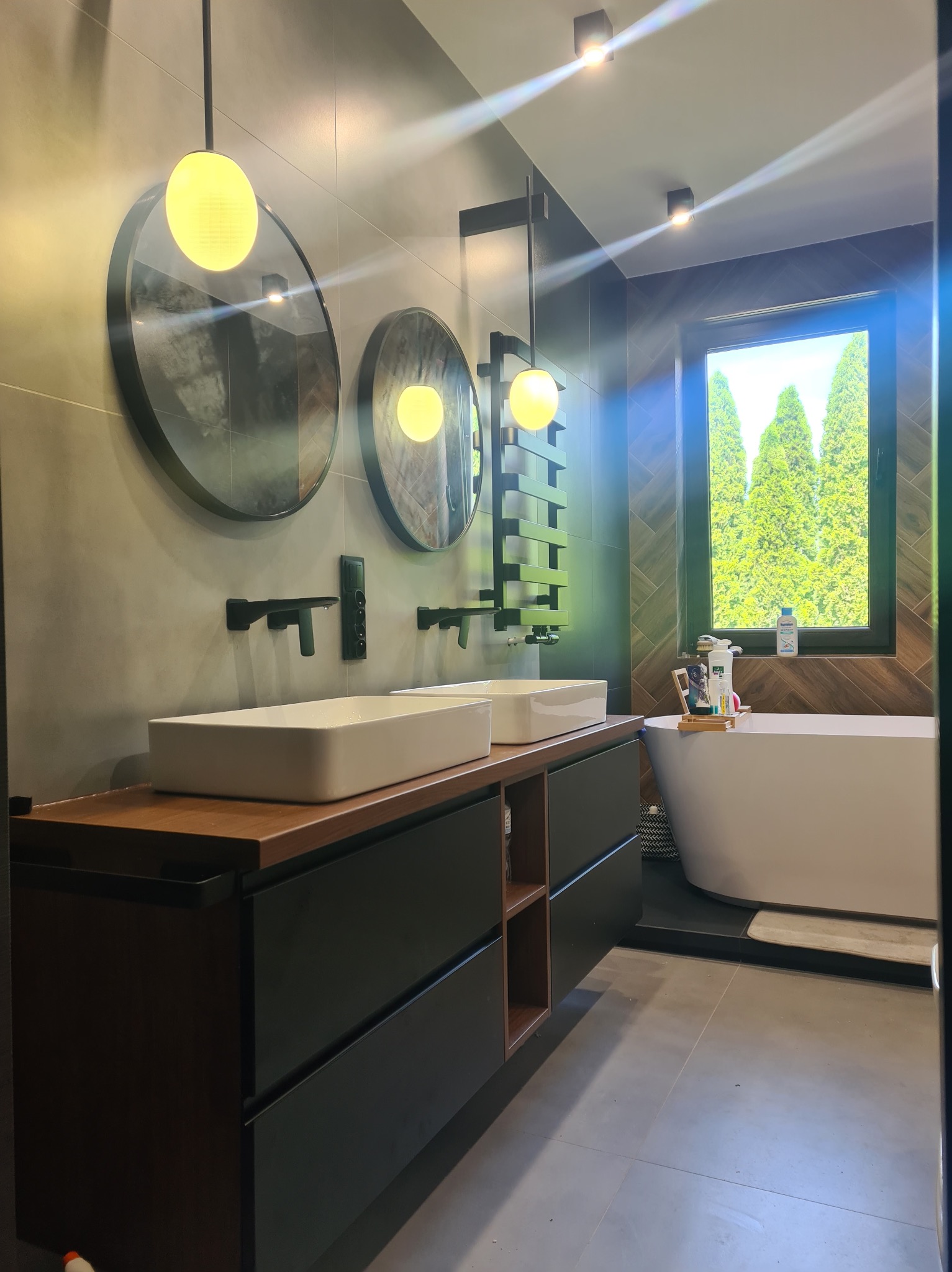 Nowoczesna łazienka z dwoma umywalkami na drewnianym blacie, okrągłymi lustrami z podświetleniem i widokiem na zieleń za oknem. Minimalistyczny design.