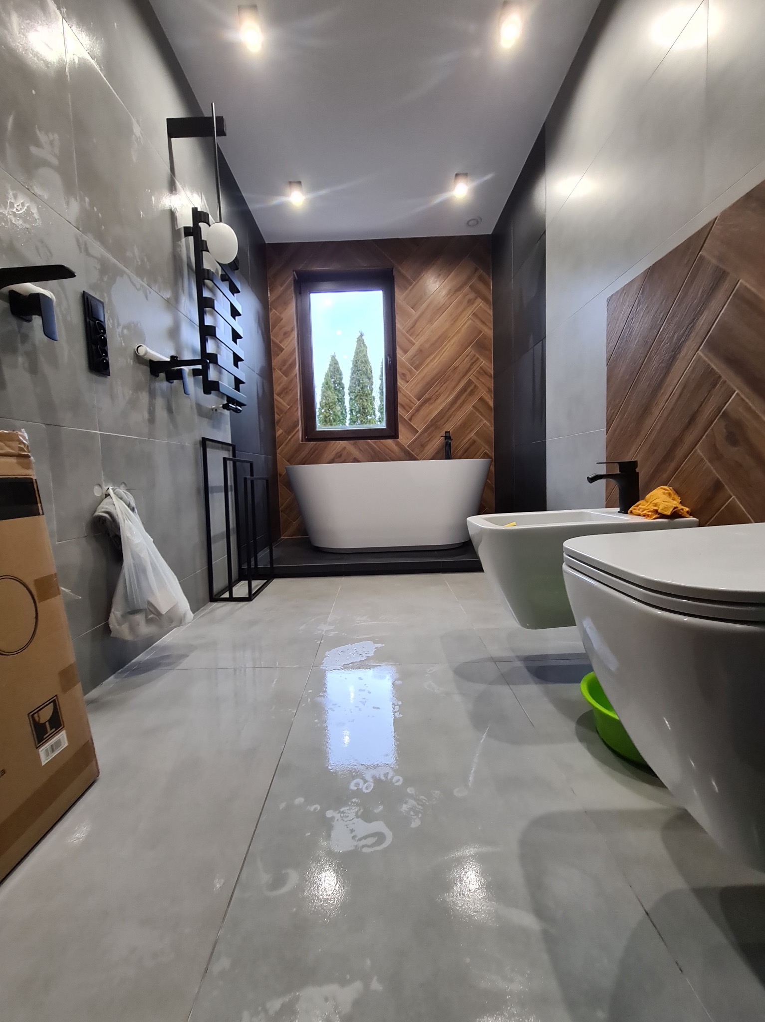 Nowoczesna łazienka z drewnianą ścianą w jodełkę, białą wanną wolnostojącą i szarą armaturą. Na podłodze widoczne ślady po remoncie, co sugeruje świeżo zakończone prace.