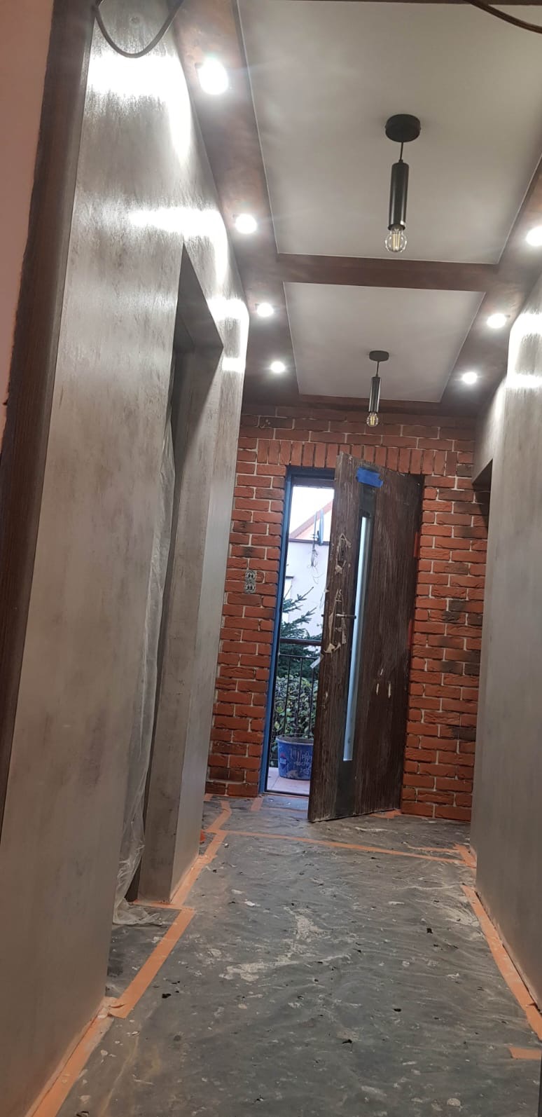 Korytarz w trakcie prac wykończeniowych: ściany z efektem betonu, ceglana ściana z otwartymi drzwiami na balkon, sufit z oświetleniem punktowym i wiszącą lampą.