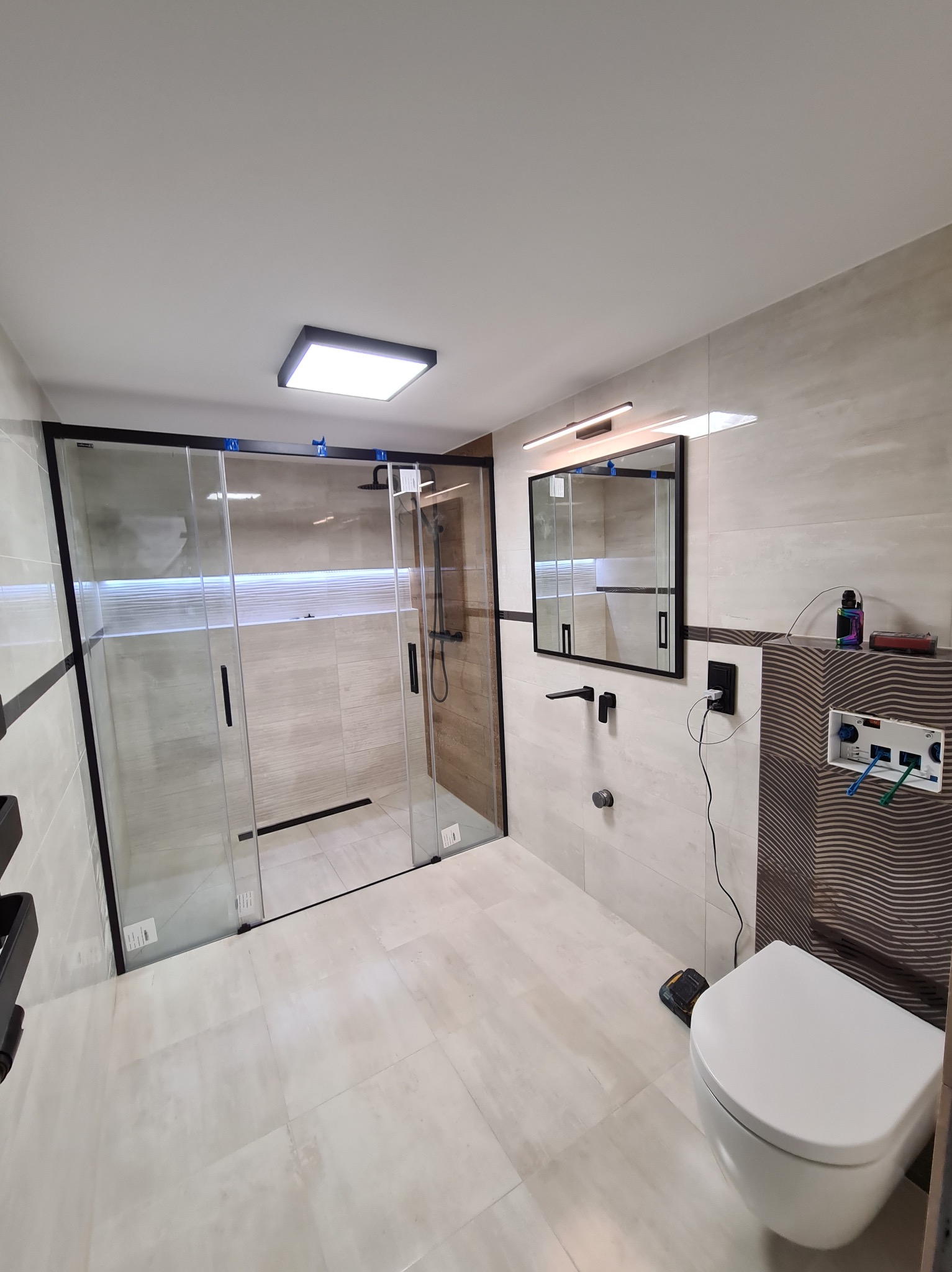 Nowoczesna łazienka z kabiną prysznicową typu walk-in, wykończona beżowymi płytkami. Czarna armatura, minimalistyczne lustro z oświetleniem i podwieszana toaleta.