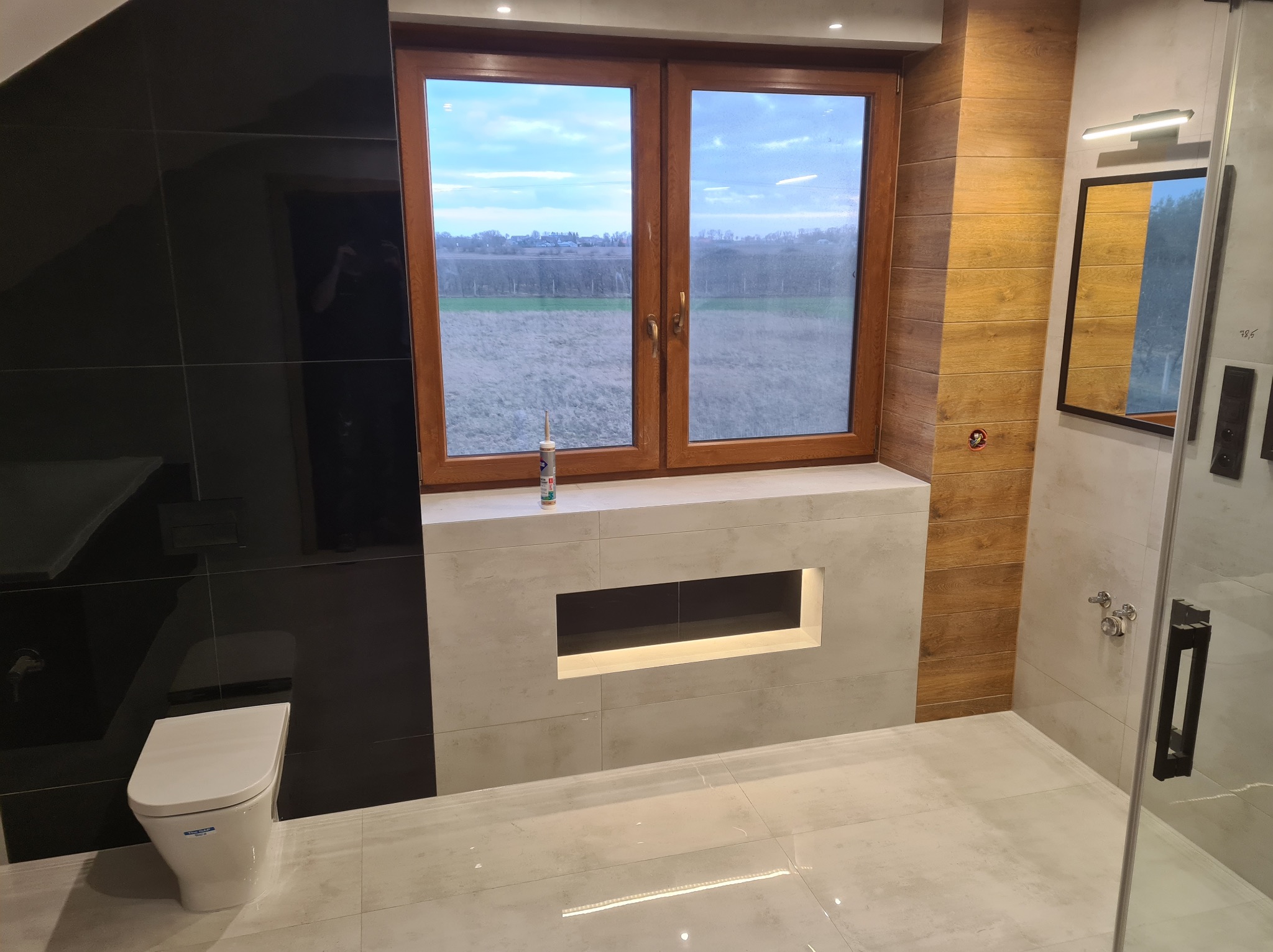 Nowoczesna łazienka z czarnymi i drewnopodobnymi płytkami, duże okno z widokiem na pole, podświetlana wnęka nad toaletą, kabina prysznicowa ze szklanymi drzwiami.