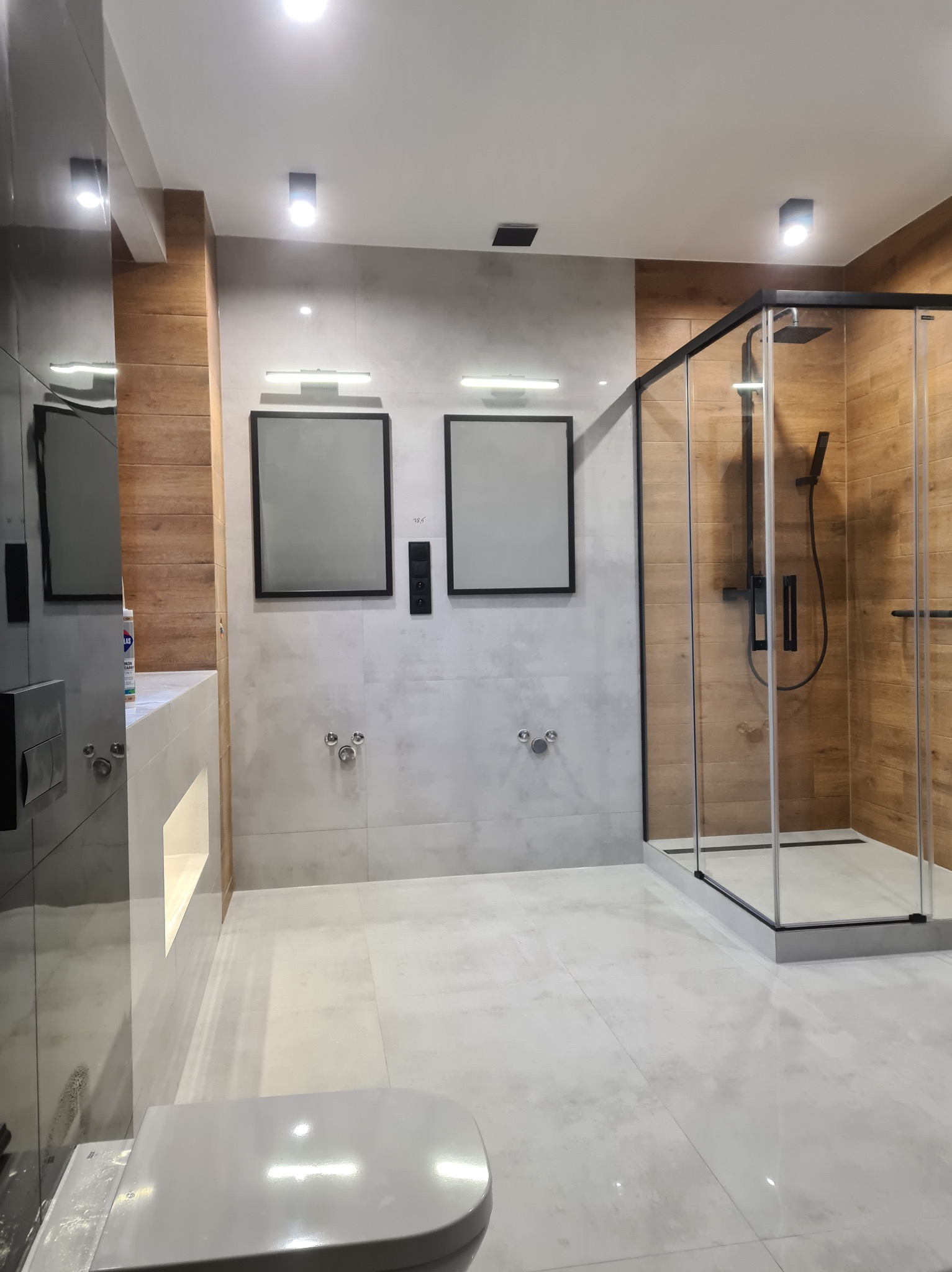 Wykończona łazienka z drewnopodobnymi płytkami w kabinie prysznicowej typu walk-in, szare płytki na ścianie i podłodze, minimalistyczne lustra w czarnych ramach.