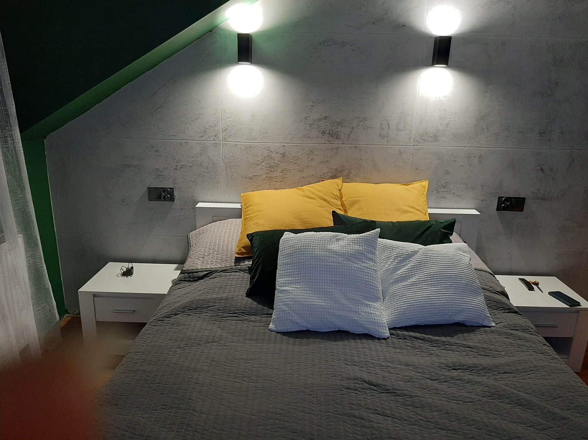 Nowoczesna sypialnia z szarą ścianą imitującą beton, oświetlona lampami ściennymi. Na łóżku żółte, zielone i białe poduszki. Widoczne białe szafki nocne.