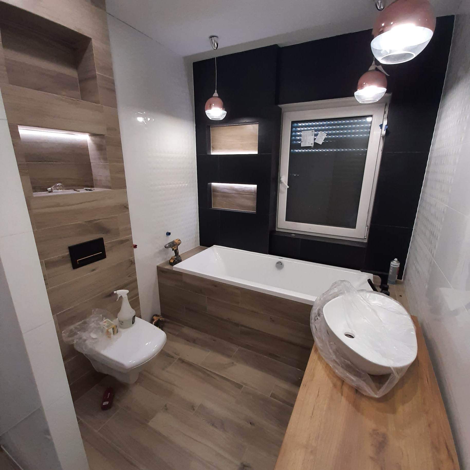 Nowoczesna łazienka z drewnopodobnymi płytkami, czarnymi akcentami i trzema okrągłymi lampami. Umywalka i toaleta zakryte folią ochronną, narzędzia obok wanny.