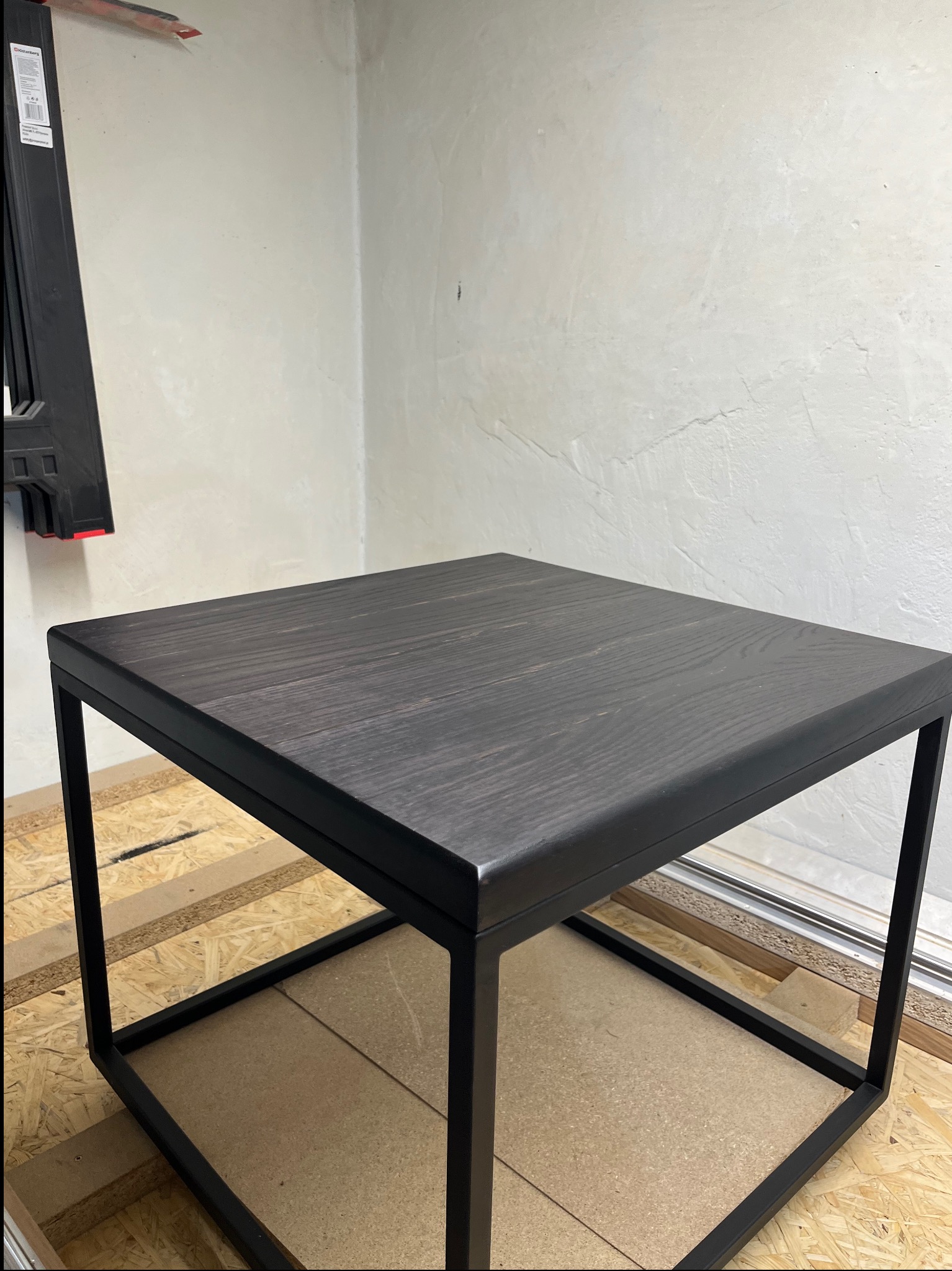 Kwadratowy stolik kawowy z blatem z ciemnego drewna i czarną, metalową ramą na tle surowej ściany i podłogi z płyt OSB.