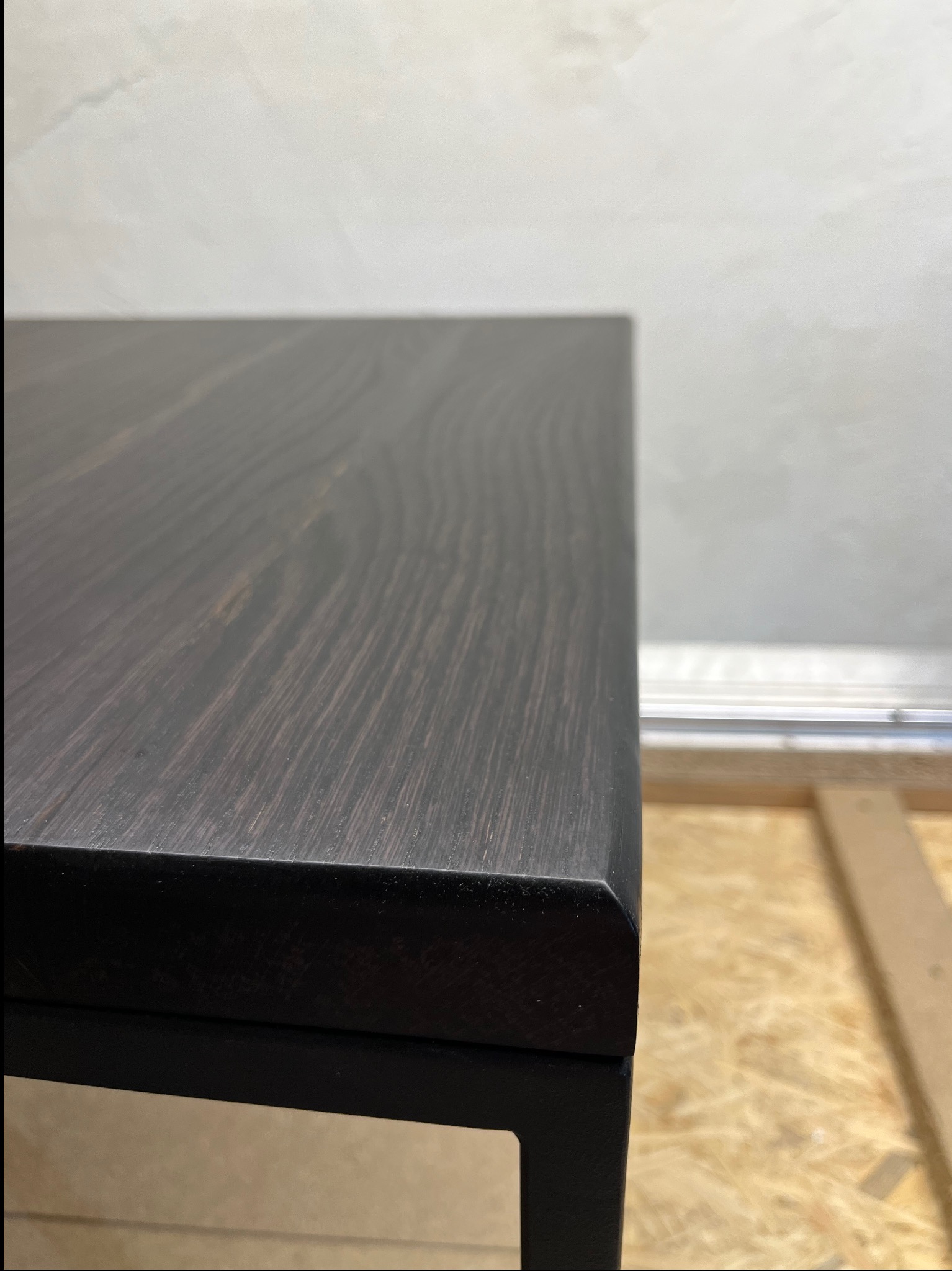 Fragment blatu z ciemnego drewna na metalowej ramie, widoczny na tle ściany i elementów konstrukcyjnych.