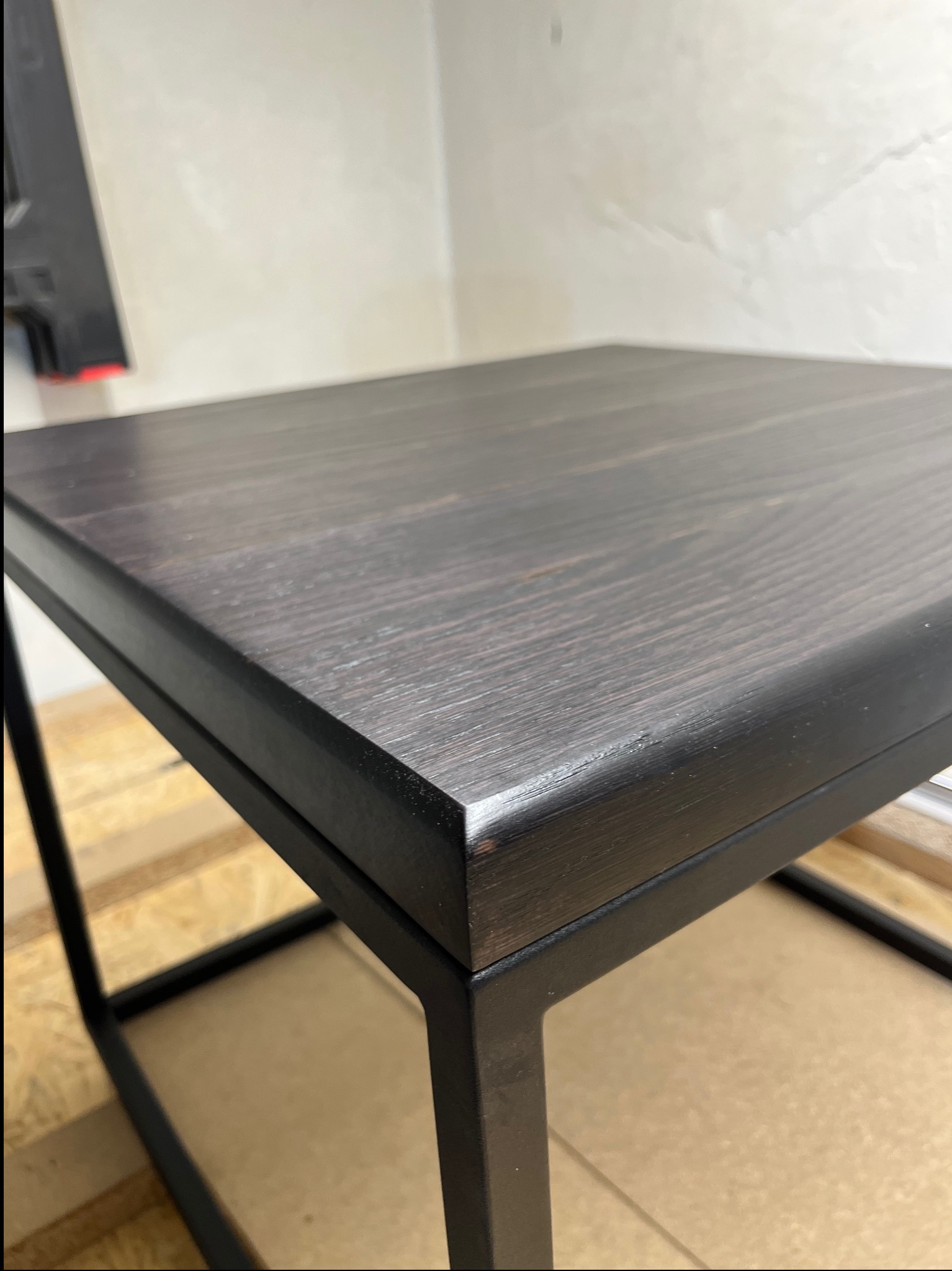 Ciemny, lakierowany blat drewniany z widocznym usłojeniem, osadzony na metalowej, czarnej ramie. Minimalistyczny design, surowe tło ściany.