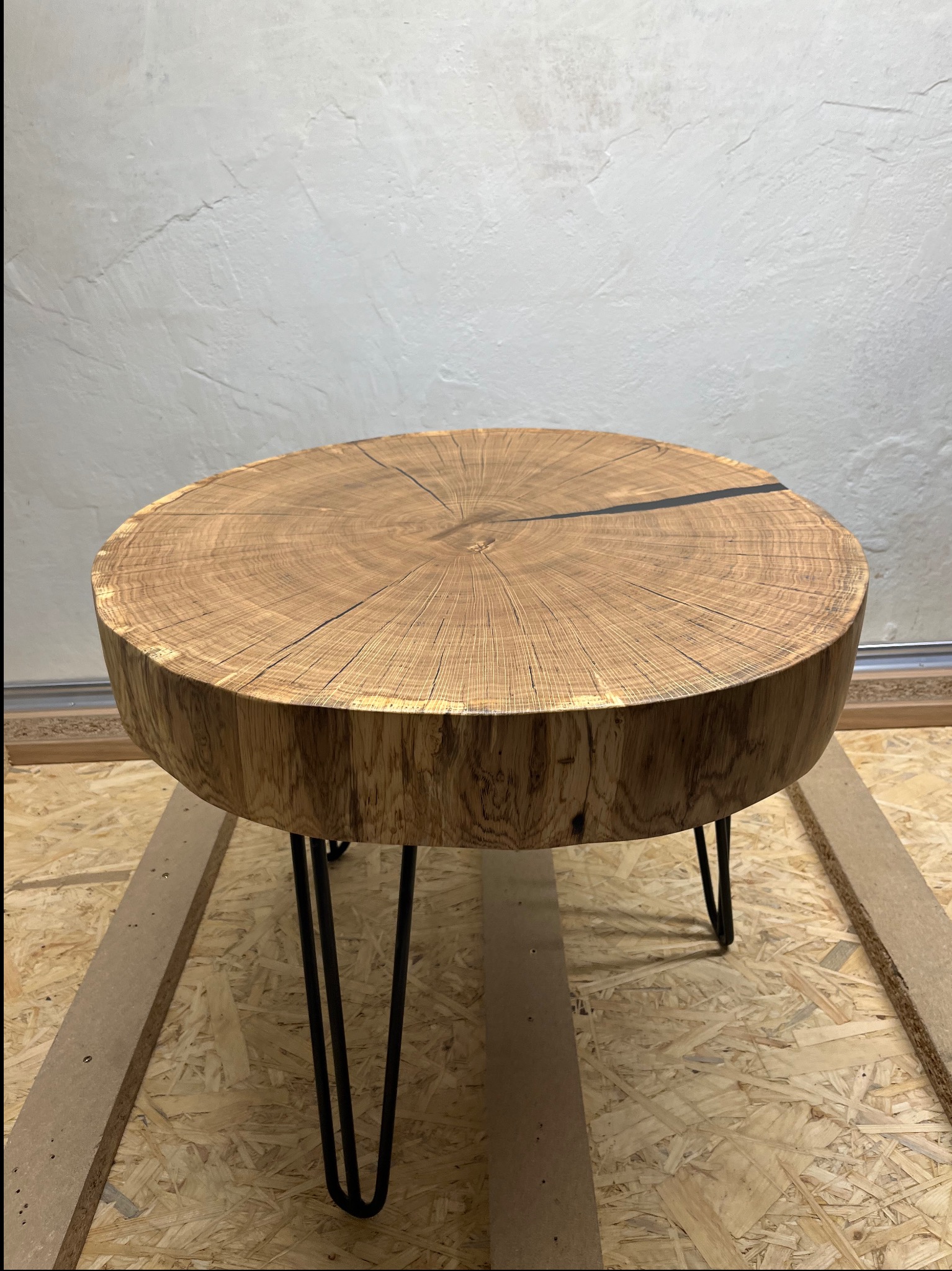 Okrągły stolik kawowy z blatem z litego drewna, ukazujący naturalne słoje i czarne wypełnienie pęknięcia, osadzony na trzech czarnych, metalowych nogach typu hairpin, na tle surowej ściany.