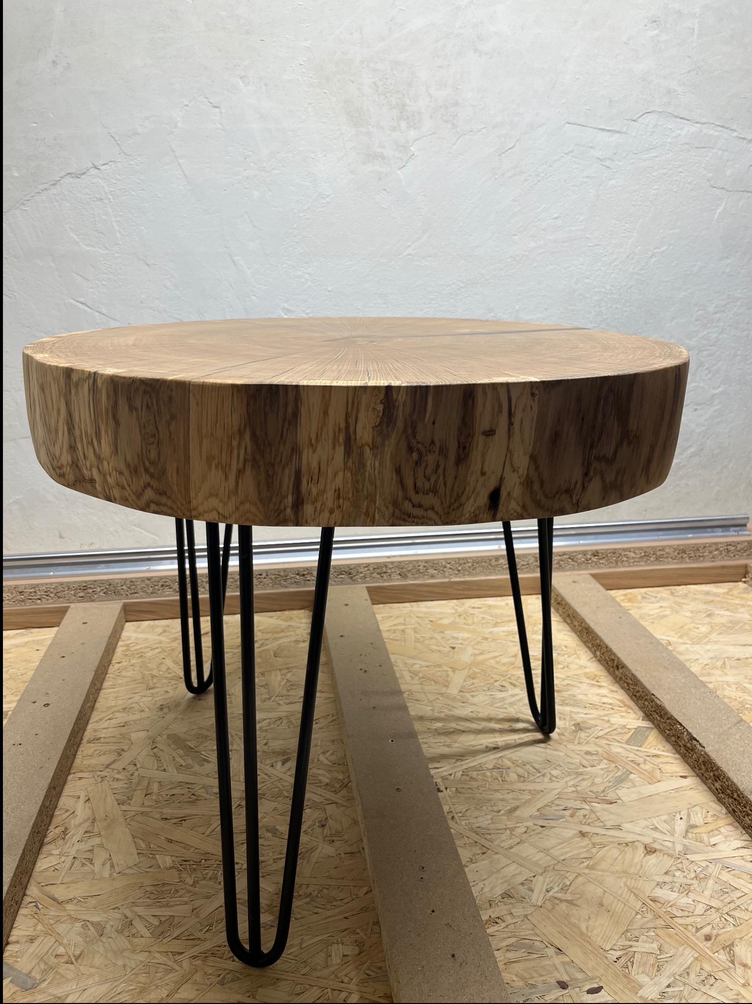 Okrągły stolik kawowy z grubego plastra drewna o widocznych słojach, osadzony na trzech czarnych, metalowych nogach typu hairpin, umieszczony w warsztacie na płycie OSB.
