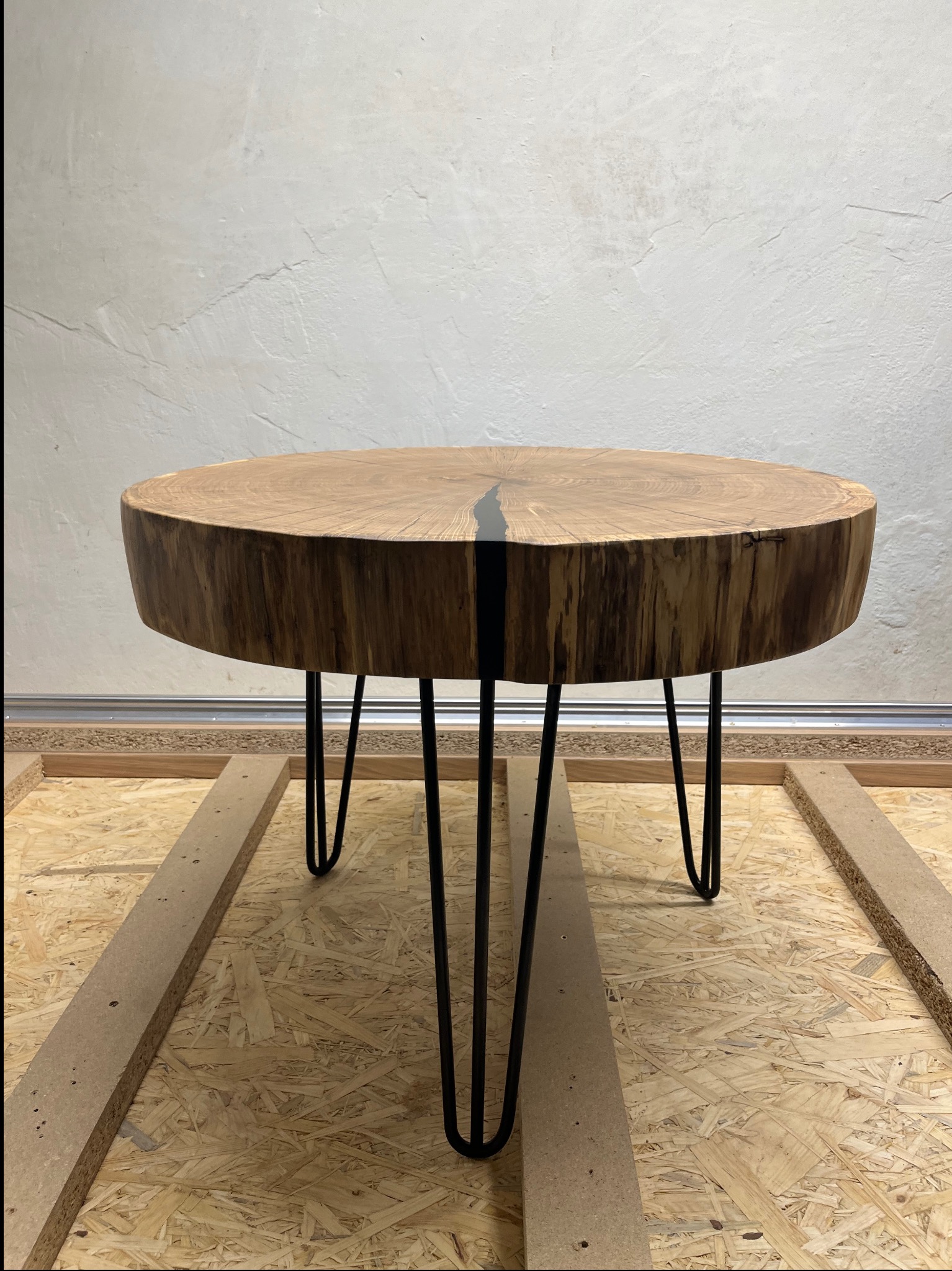 Okrągły stolik kawowy z blatu z litego drewna z żywicą epoksydową, umieszczony na metalowych nóżkach typu hairpin, na tle surowej ściany.