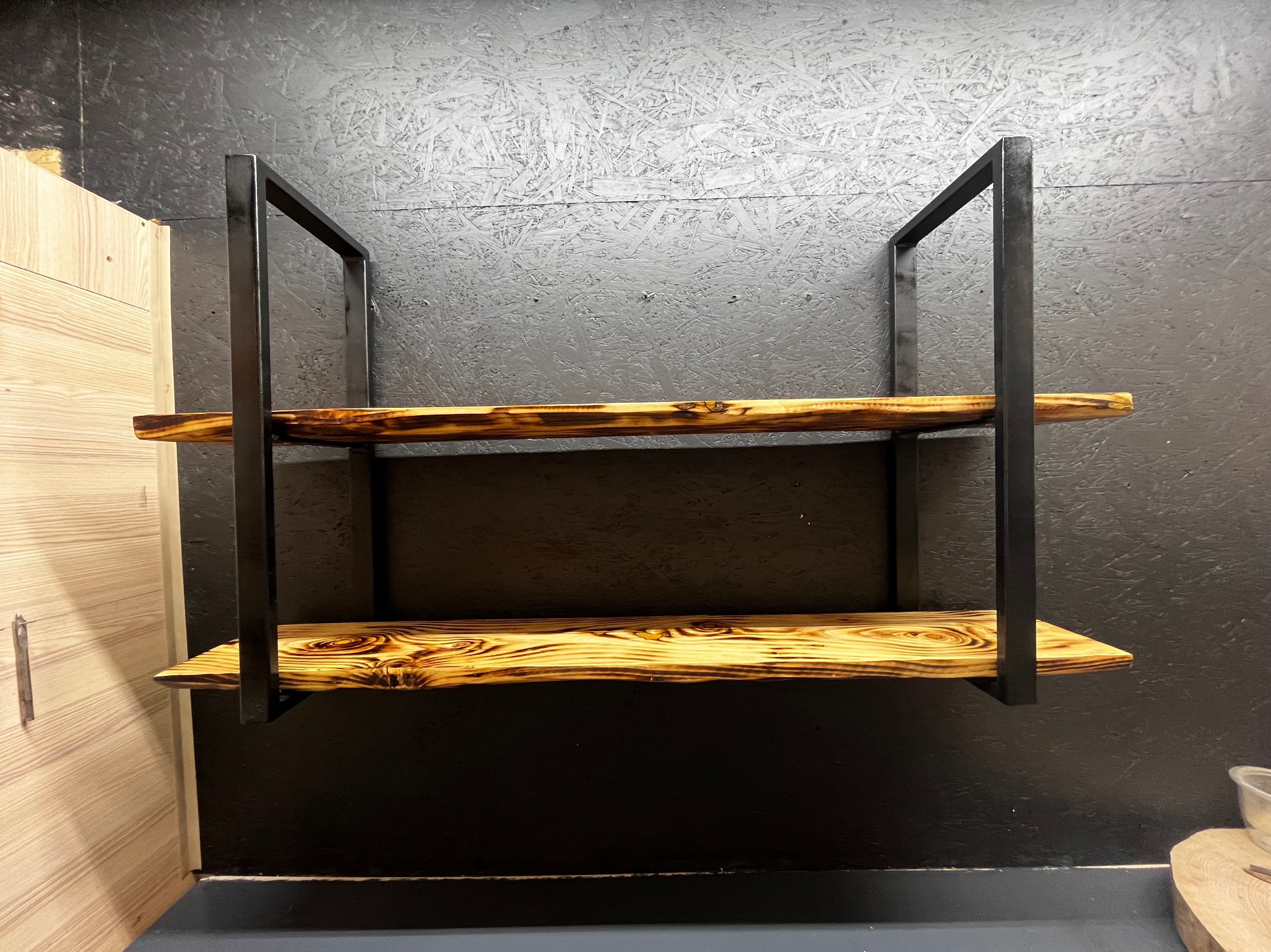 Dwie półki z opalanych desek, osadzone w metalowej, czarnej ramie, zamontowane na ciemnej ścianie z płyty OSB.