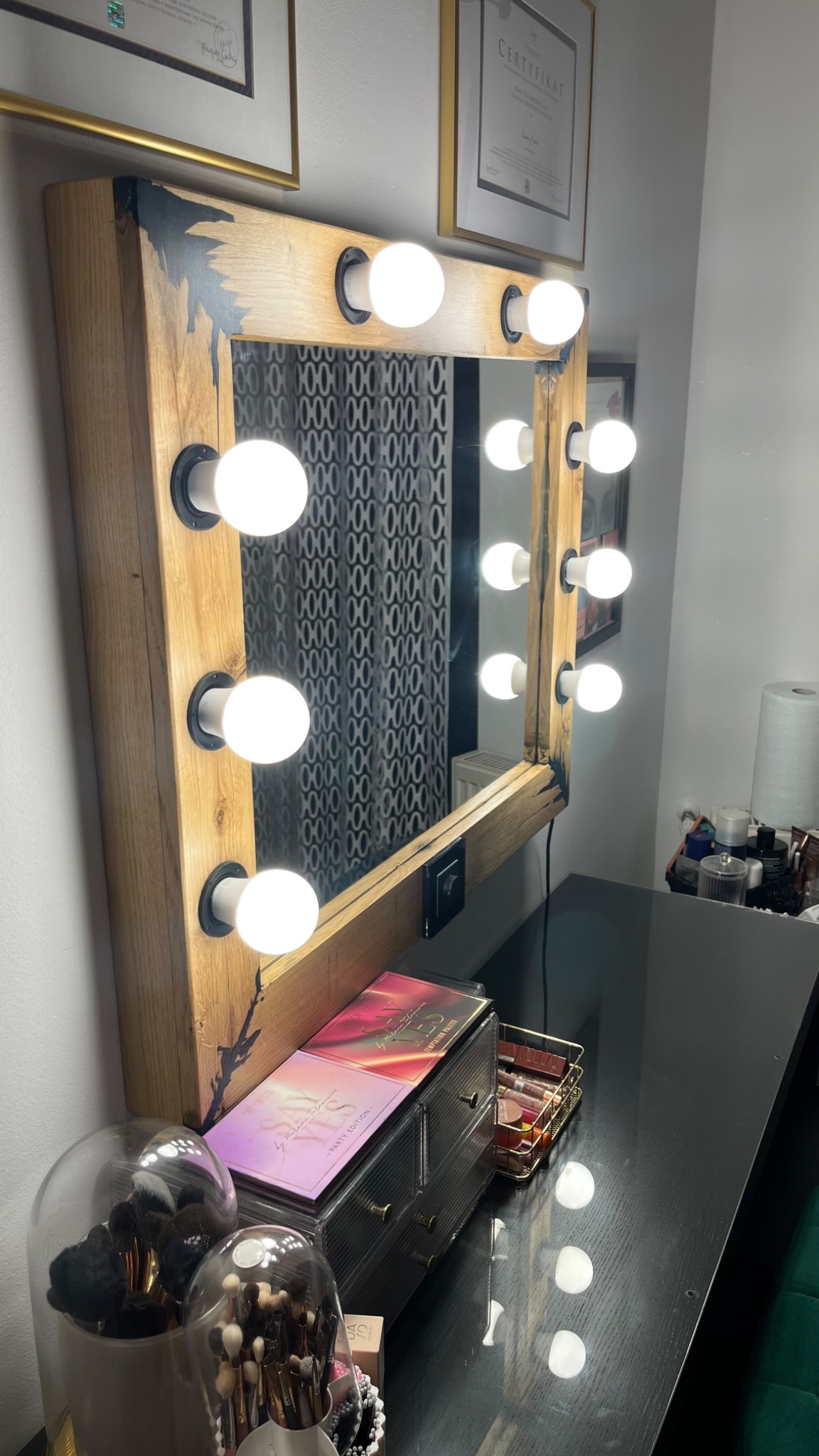Lustro do makijażu z drewnianą ramą i oświetleniem żarówkowym, stojące na czarnym blacie, na którym widoczne są kosmetyki i akcesoria do makijażu.