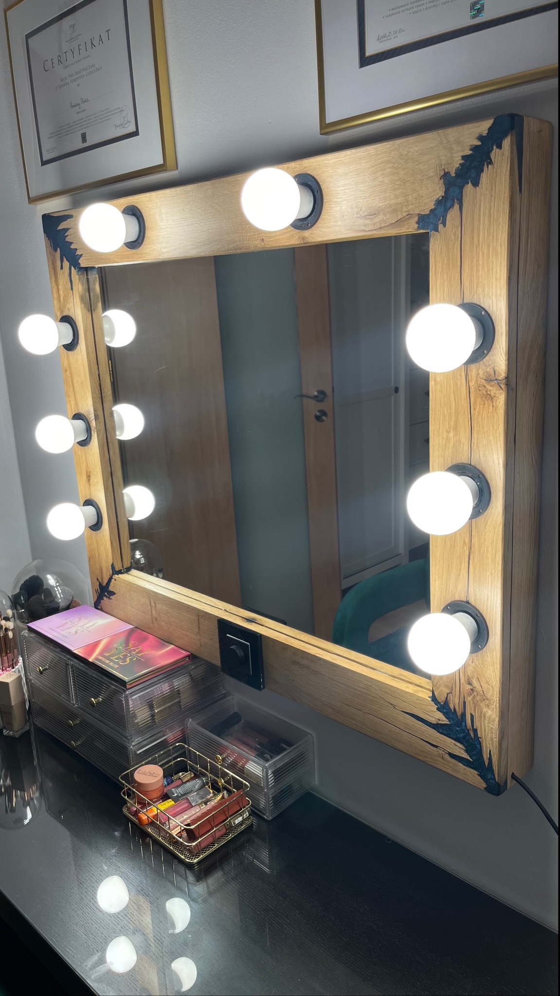 Rustykalne lustro do makijażu z drewnianą ramą, oświetlone żarówkami, stojące na czarnym blacie toaletki, obok kosmetyki i certyfikat w tle.
