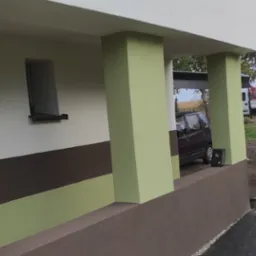 Świeżo pomalowana elewacja domu z kolumnami w odcieniach zieleni i brązu, widoczne okno, zaparkowany samochód i ciężarówka w tle.