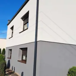 Dwukondygnacyjny dom z nową, dwukolorową elewacją: biała góra i szary dół. Widoczne okna z brązowymi ramami i szarymi roletami oraz ciemna rynna.